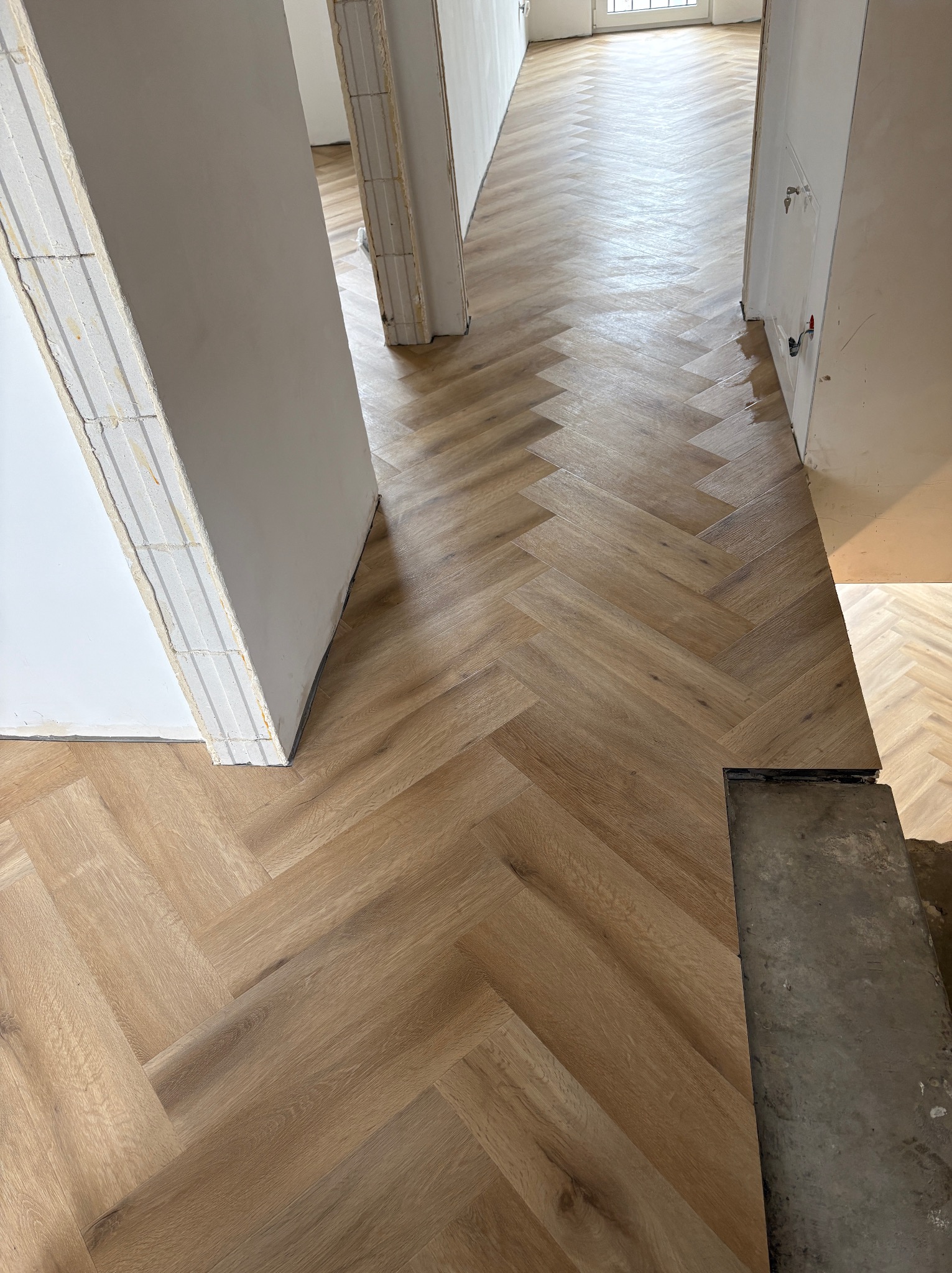 Ułożona podłoga z paneli w jodełkę, widok z góry, jasne panele, surowe ściany w tle, fragment progu. Podłoga po remoncie.