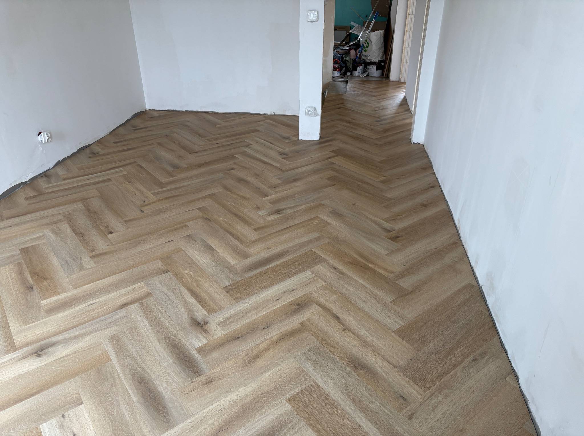 Ułożona podłoga z paneli w jodełkę francuską, jasne drewno, widoczne białe ściany i fragment kolumny. W tle otwarte przejście do innego pomieszczenia.