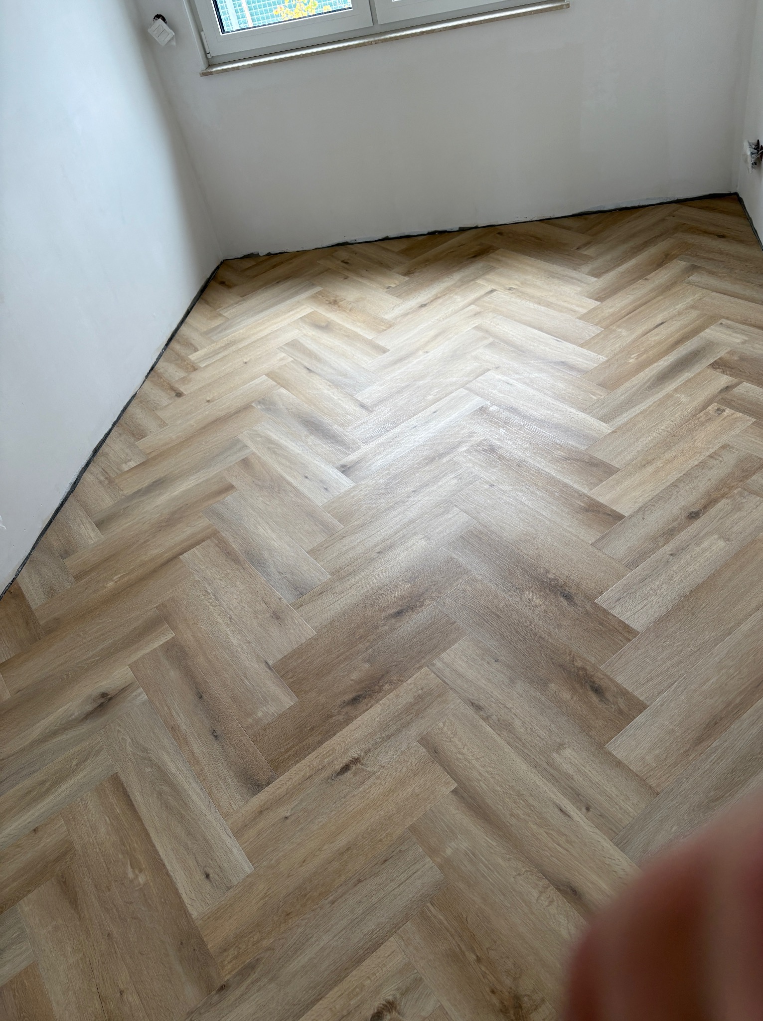 Jasne panele ułożone w jodełkę w pokoju z białymi ścianami i oknem. Widoczny fragment dłoni w prawym dolnym rogu kadru. Nowoczesne wykończenie podłogi.