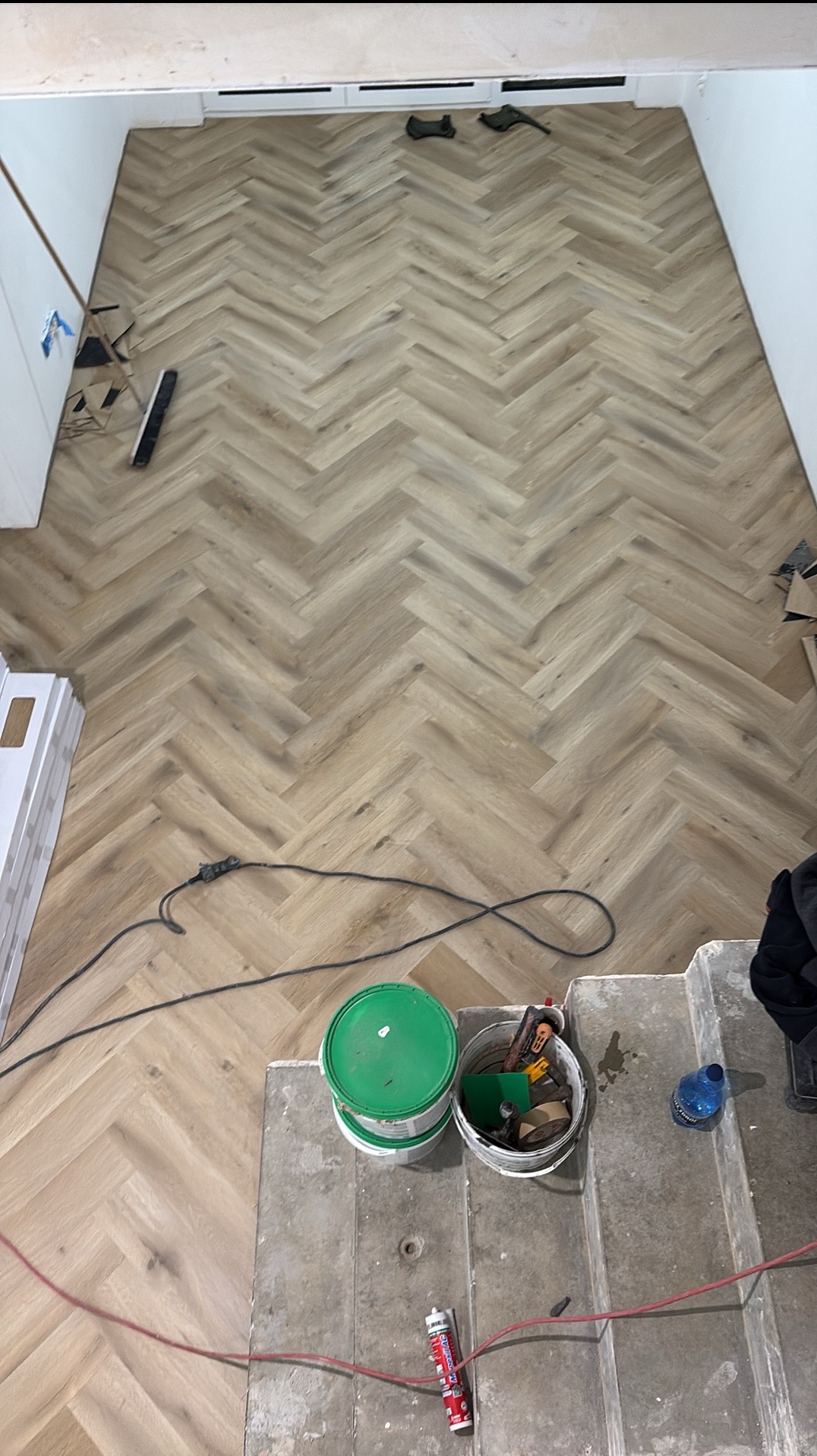 Ukończona podłoga z paneli ułożonych w jodełkę, widok z góry. Na pierwszym planie narzędzia i materiały budowlane. Jasne panele, naturalne światło.