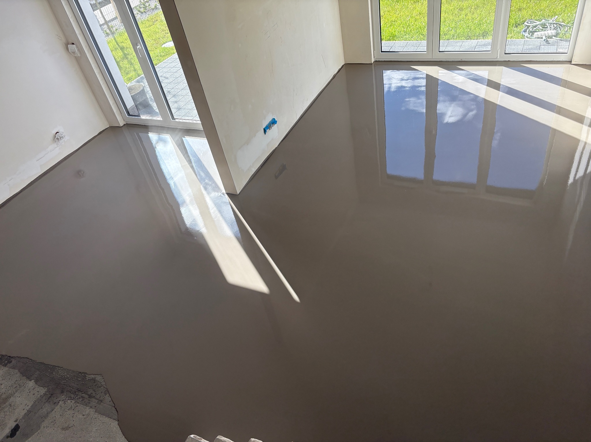 Wylany, gładki podkład podłogowy w jasnym wnętrzu z dużymi oknami, odbijającymi światło słoneczne. Widoczne fragmenty schodów i ściany z instalacją.