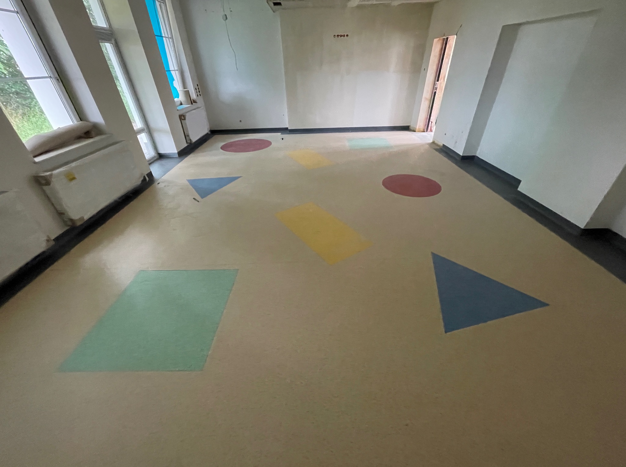 Jasna wykładzina w pokoju z kolorowymi figurami geometrycznymi: koła, trójkąty i prostokąty. Widok na okno i grzejnik.