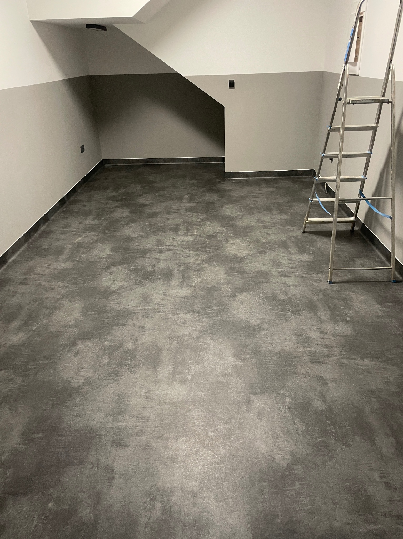 Szara, matowa podłoga z wykładziny w pomieszczeniu z drabinką. Wykończenie ścian w dwóch odcieniach szarości, z ciemniejszym pasem na dole. Minimalistyczny design.