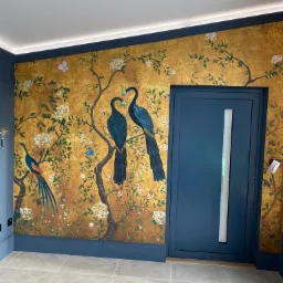 Dekoracyjna tapeta z motywem ptaków i kwiatów na złotej ścianie, dopasowana kolorystycznie do ciemnoniebieskiej stolarki drzwiowej i listew przypodłogowych w korytarzu z płytkami podłogowymi...