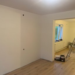 Bartłomiej Sawicki - Świeżo pomalowane na biało wnętrze z drewnianą podłogą. Widoczne puszki farb, drabina i lampa malarska. Dwa otwory na gniazdka elektryczne na ścianie.