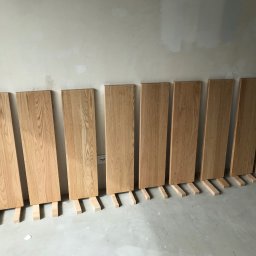 Bartłomiej Sawicki - Dębowe panele ułożone pionowo wzdłuż ściany na drewnianych podkładkach, przygotowane do montażu. Widoczna surowa ściana i podłoga w trakcie wykończenia.