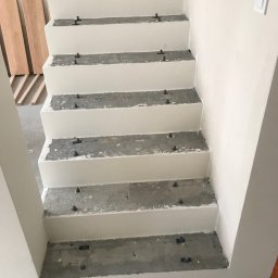 Bartłomiej Sawicki - Betonowe schody w trakcie wykańczania, z widocznymi elementami montażowymi i przygotowanymi drewnianymi deskami obok. Świeżo pomalowane ściany i włącznik światła.