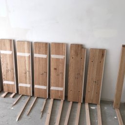 Bartłomiej Sawicki - Drewniane panele z numeracją, przygotowane do montażu przy ścianie. Widoczne listewki dystansowe na podłodze. Surowe wykończenie wnętrza, etap prac wykończeniowych.