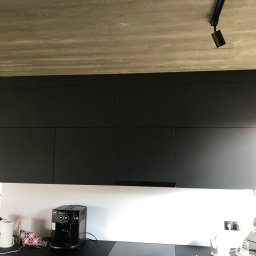 Bartłomiej Sawicki - Czarne, matowe szafki kuchenne wiszące na białej ścianie, z widocznym betonowym sufitem i oświetleniem punktowym. Na blacie ekspres do kawy i narzędzia.