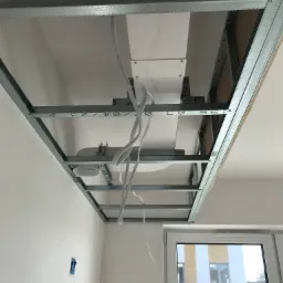 Metalowa konstrukcja sufitu podwieszanego z płyt kartonowo-gipsowych, widok od dołu, z widocznymi przewodami elektrycznymi i elementami oświetleniowymi, ściany w trakcie malowania.