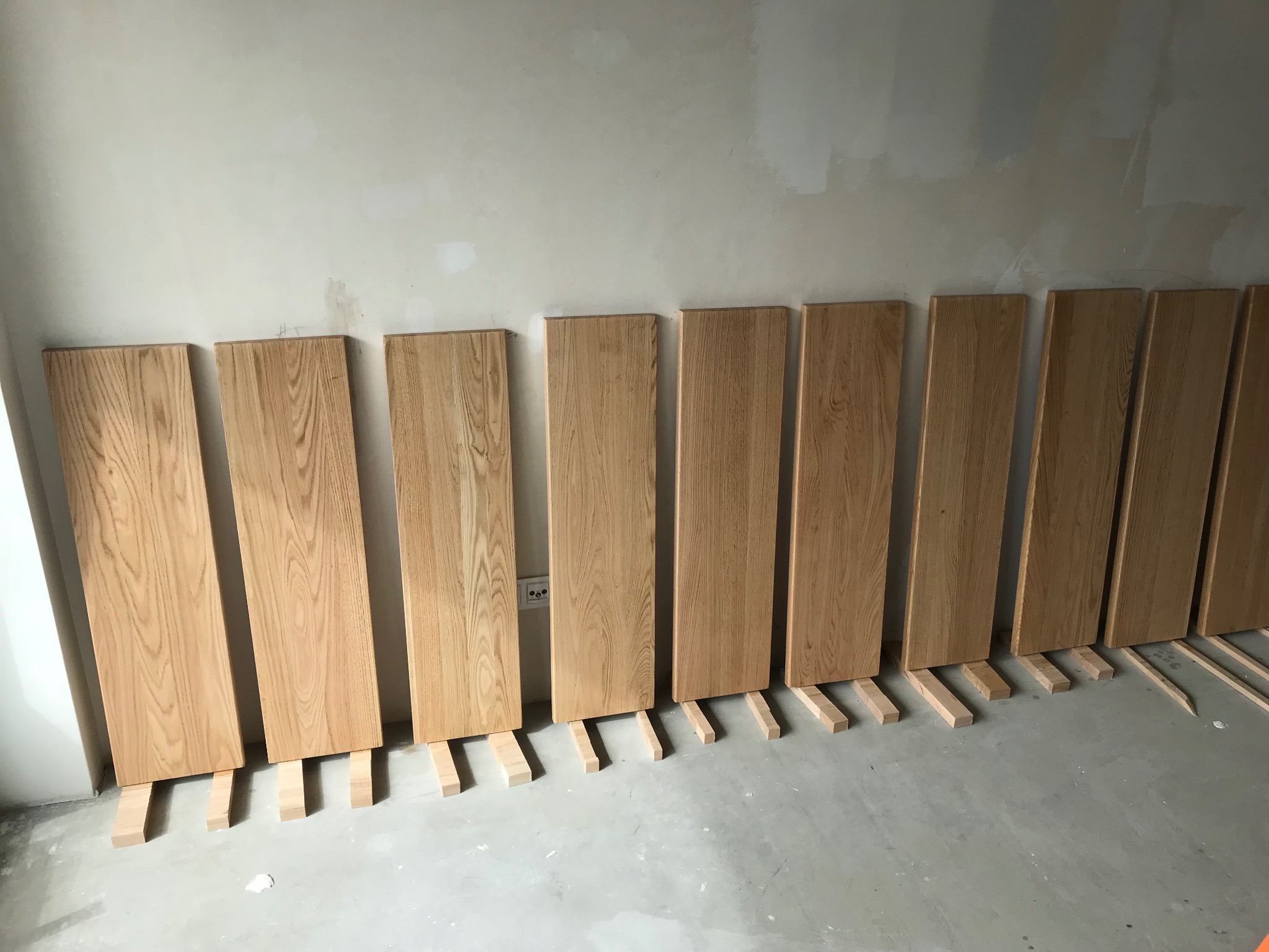 Dębowe panele ułożone pionowo wzdłuż ściany na drewnianych podkładkach, przygotowane do montażu. Widoczna surowa ściana i podłoga w trakcie wykończenia.