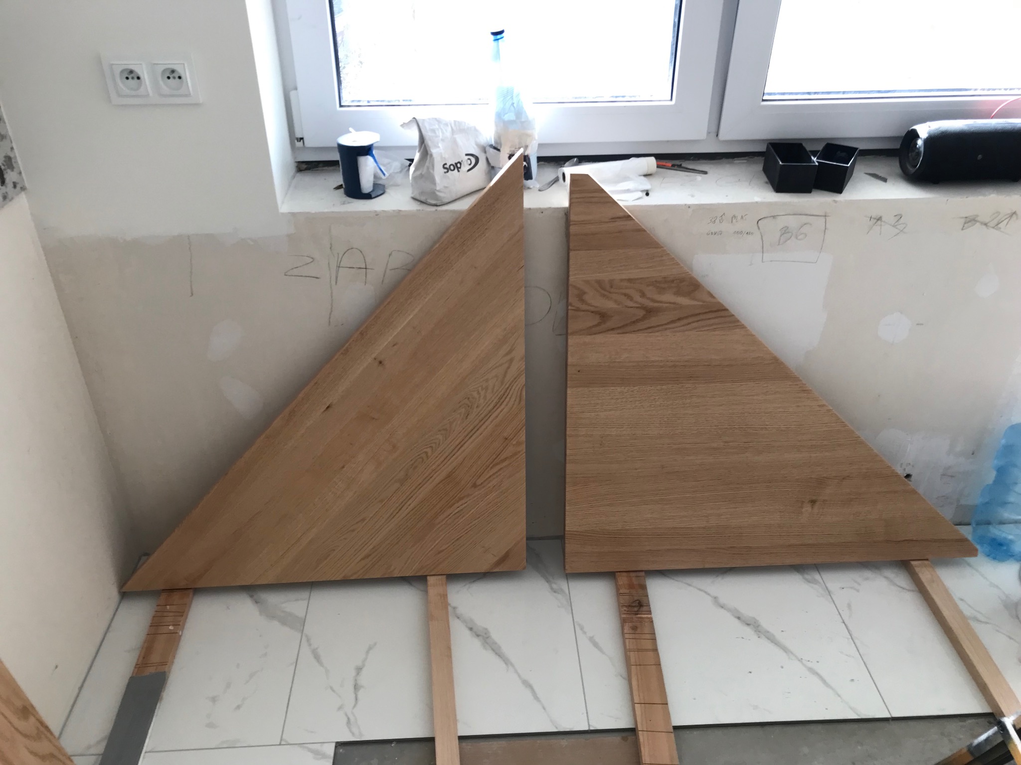 Dwa trójkątne blaty z jasnego drewna dębowego, oparte o ścianę z widocznymi śladami prac wykończeniowych, na tle okna i marmurowej podłogi. Przygotowanie do montażu.
