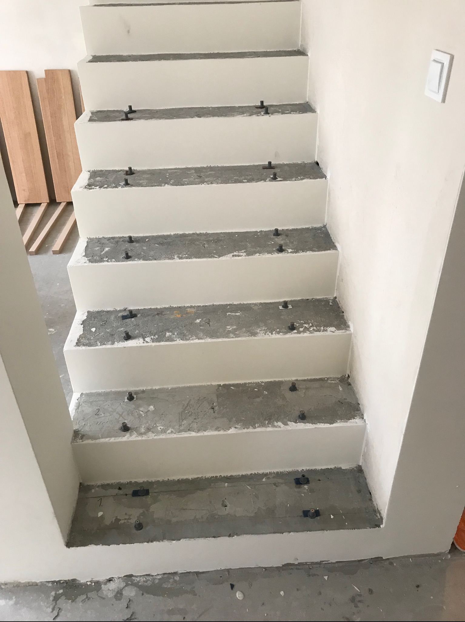 Betonowe schody w trakcie wykańczania, z widocznymi elementami montażowymi i przygotowanymi drewnianymi deskami obok. Świeżo pomalowane ściany i włącznik światła.