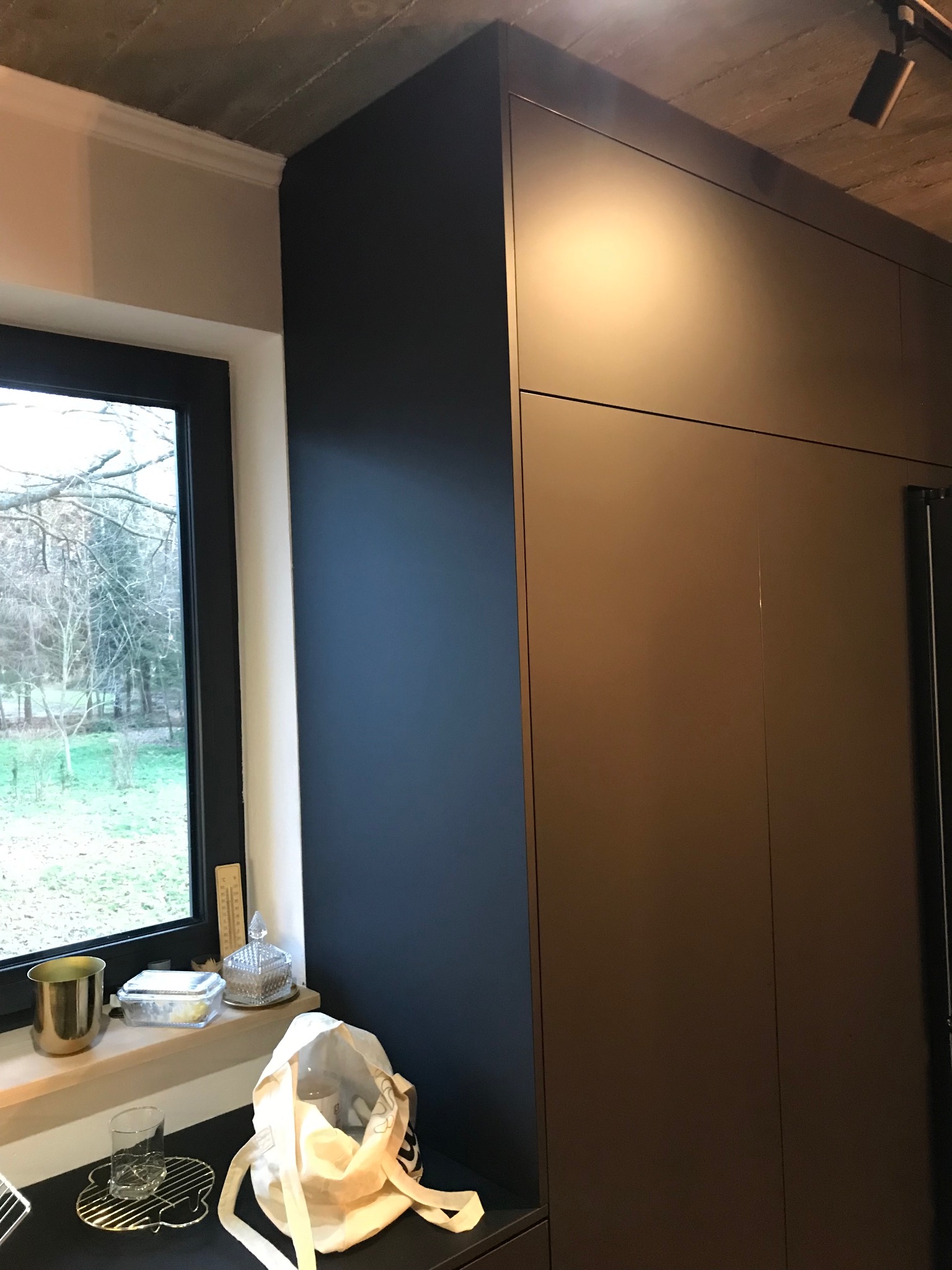 Ciemna zabudowa kuchenna na wymiar, widok z bliska. Minimalistyczny design, matowe fronty, oświetlenie punktowe. Okno z widokiem na zewnątrz. Nowoczesna aranżacja.