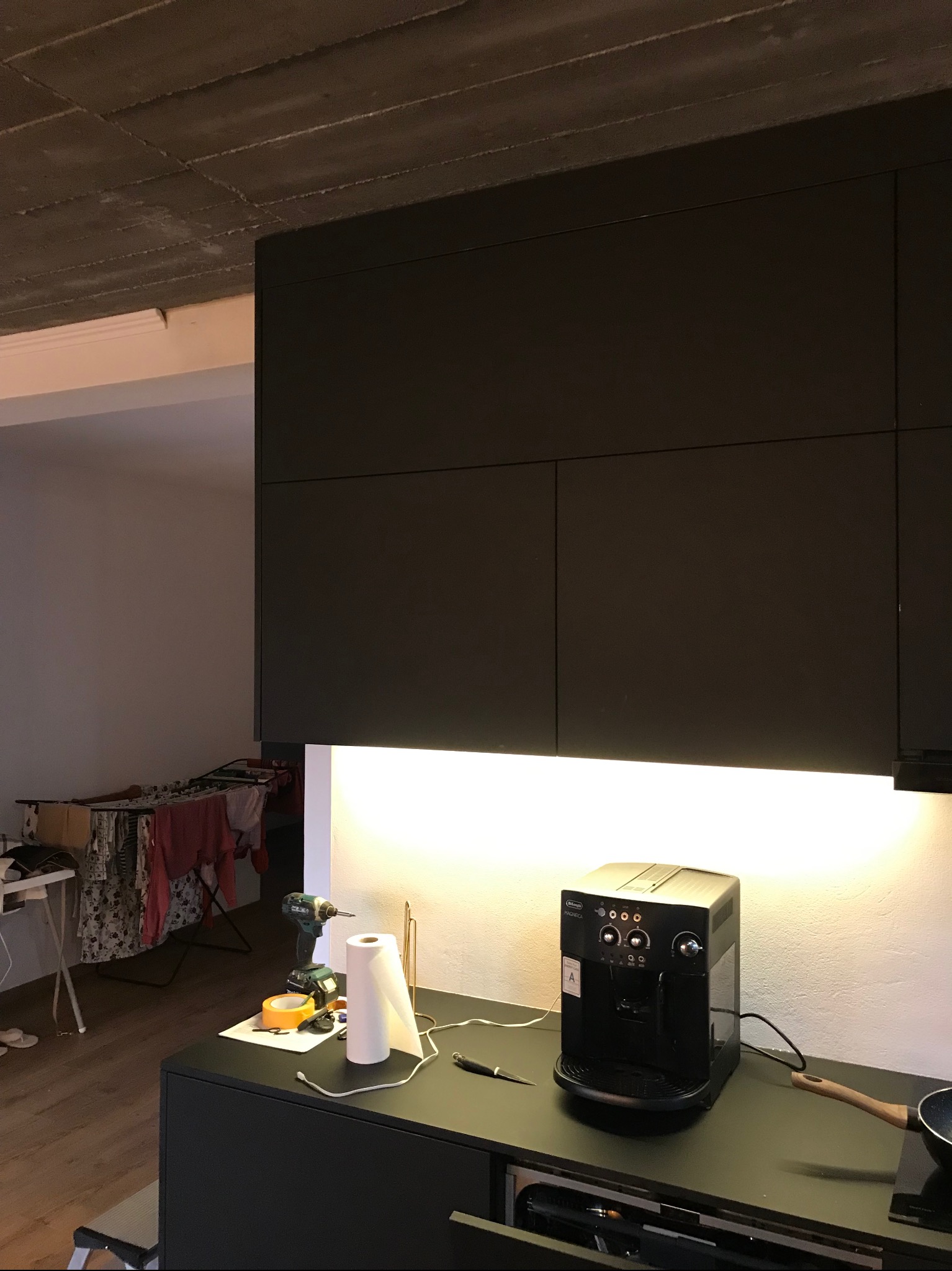 Ciemne szafki kuchenne z oświetleniem LED pod spodem, na blacie ekspres do kawy, wiertarka i akcesoria. W tle suszarka z praniem i surowy betonowy sufit.