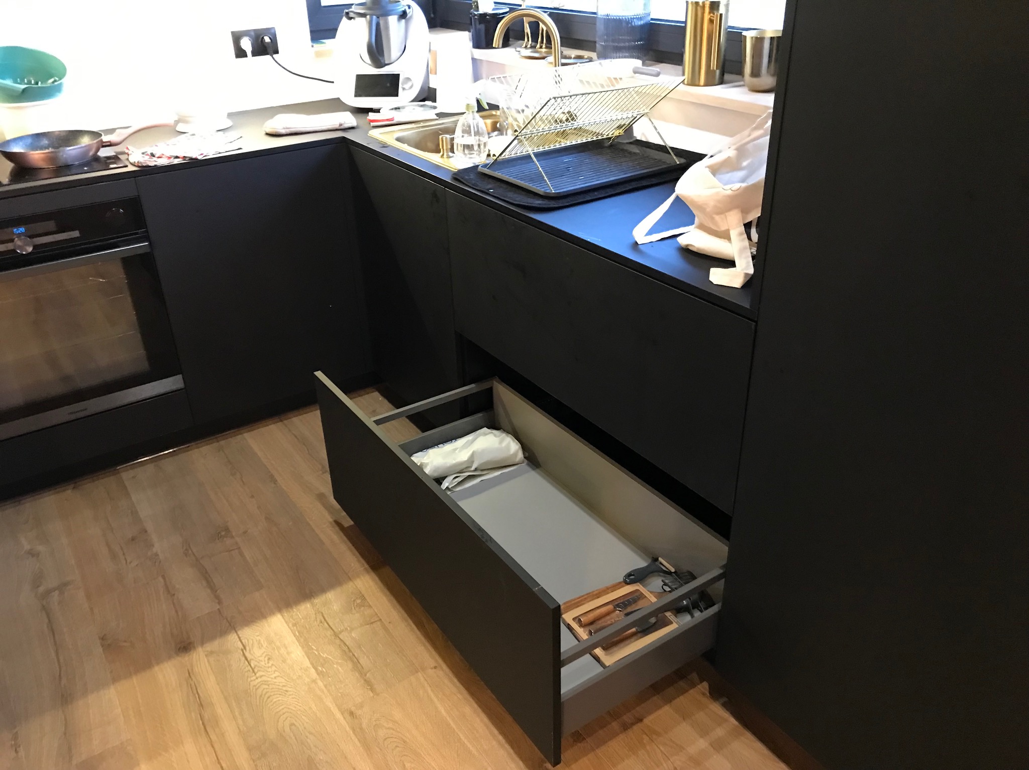 Ciemna zabudowa kuchenna z otwartą szufladą, w której widoczne są akcesoria kuchenne. Na blacie zlewozmywak i suszarka. Minimalistyczny design.