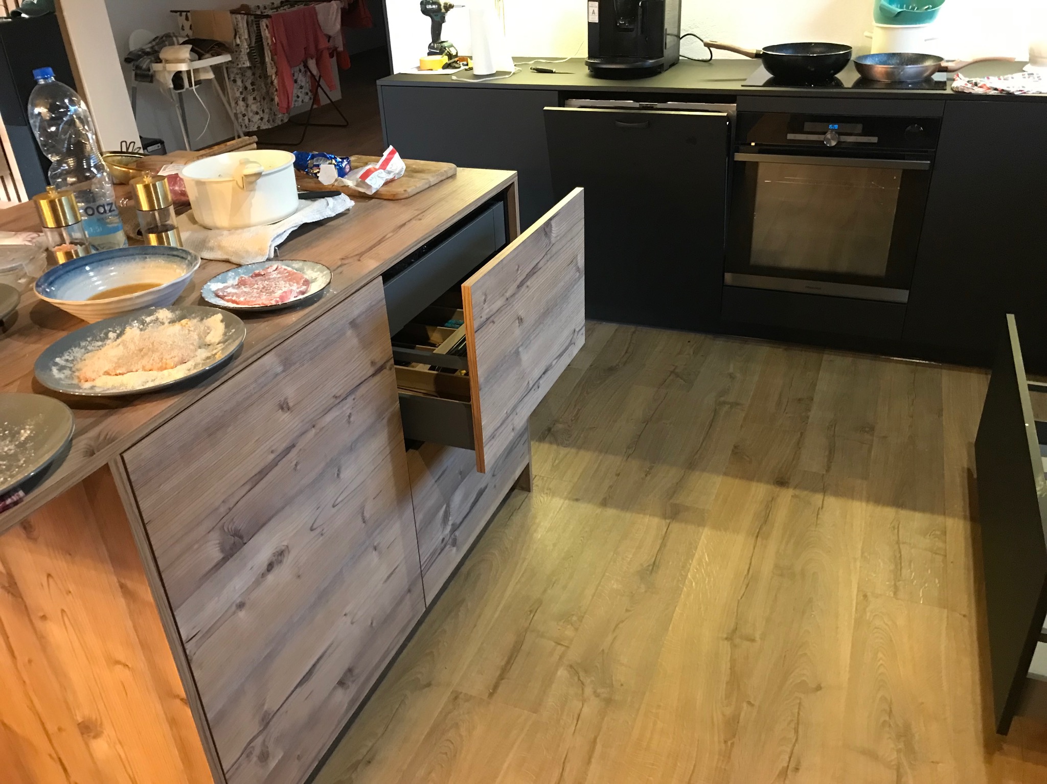 Nowoczesna kuchnia z drewnianymi frontami szafek i czarnymi elementami. Na blacie naczynia i produkty spożywcze, otwarty szuflada. Jasna podłoga.