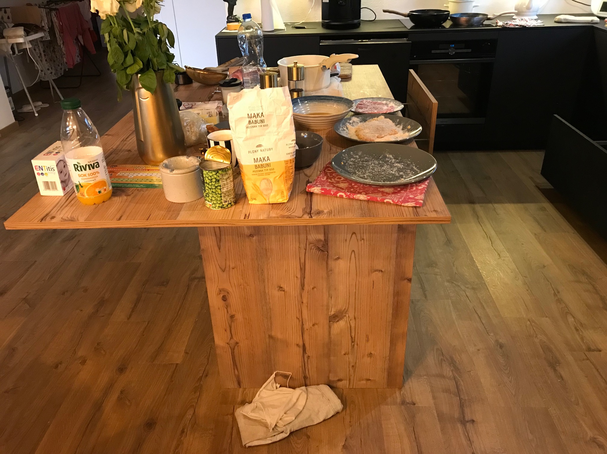 Przygotowanie posiłku na drewnianym stole w kuchni w Krosno. Widoczne produkty spożywcze, naczynia i kwiaty. Ujęcie z perspektywy osoby stojącej.