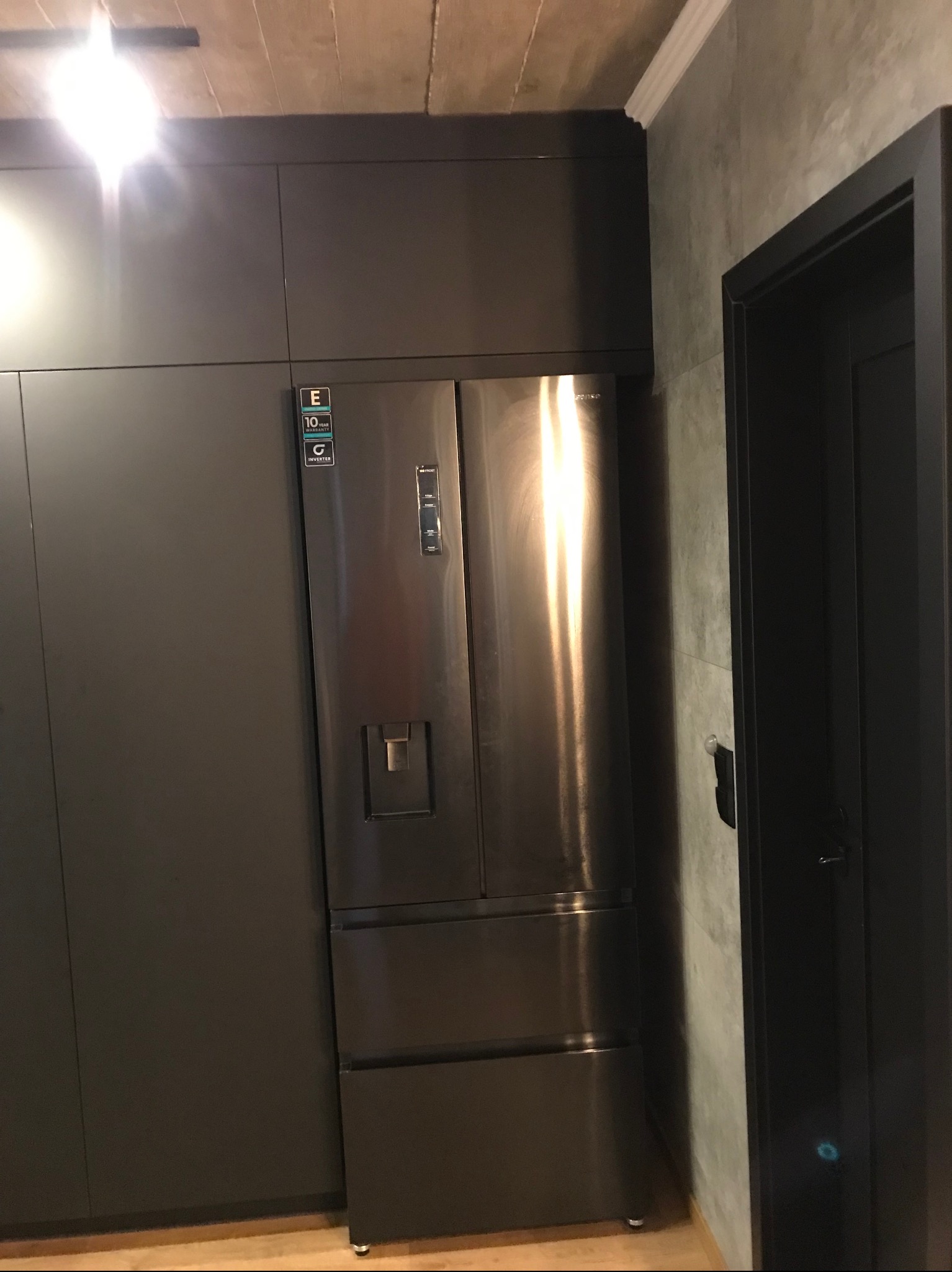 Nowoczesna kuchnia z zabudową w odcieniach szarości, zintegrowana z lodówką side-by-side. Minimalistyczny design, surowy beton na suficie, eleganckie wykończenie.