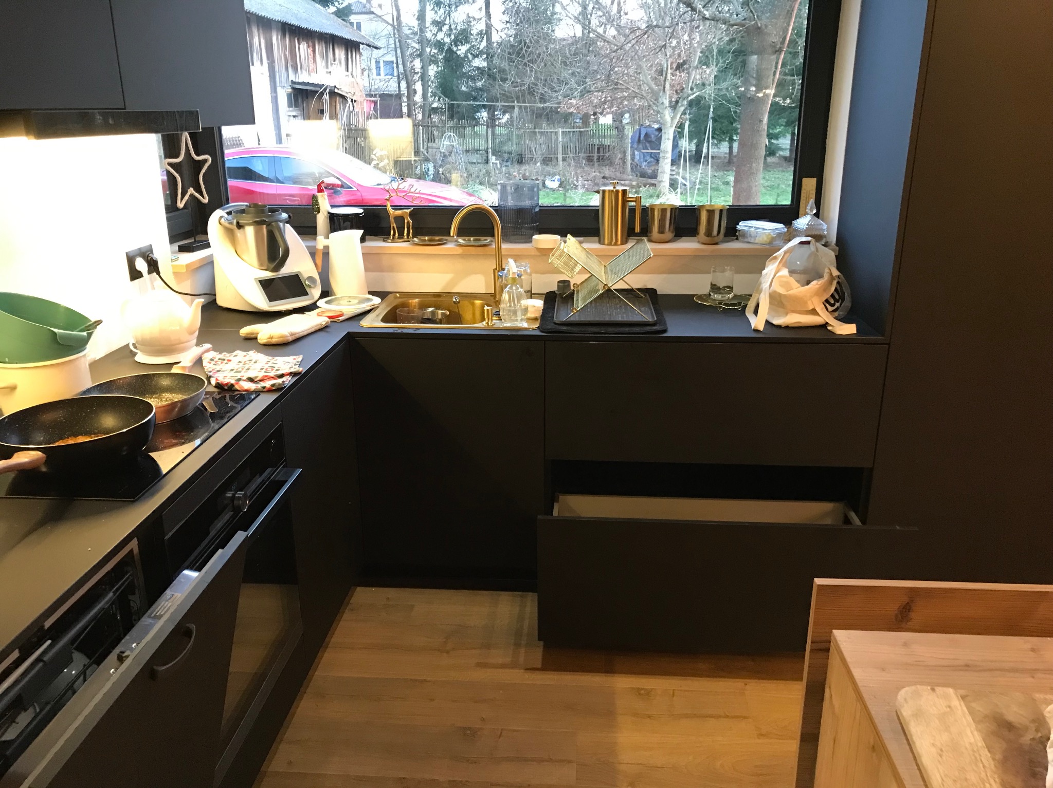 Nowoczesna, czarna kuchnia z drewnianą podłogą i złotą armaturą. Na blacie widoczne akcesoria kuchenne i okno z widokiem na zewnątrz. Otwarta szuflada.