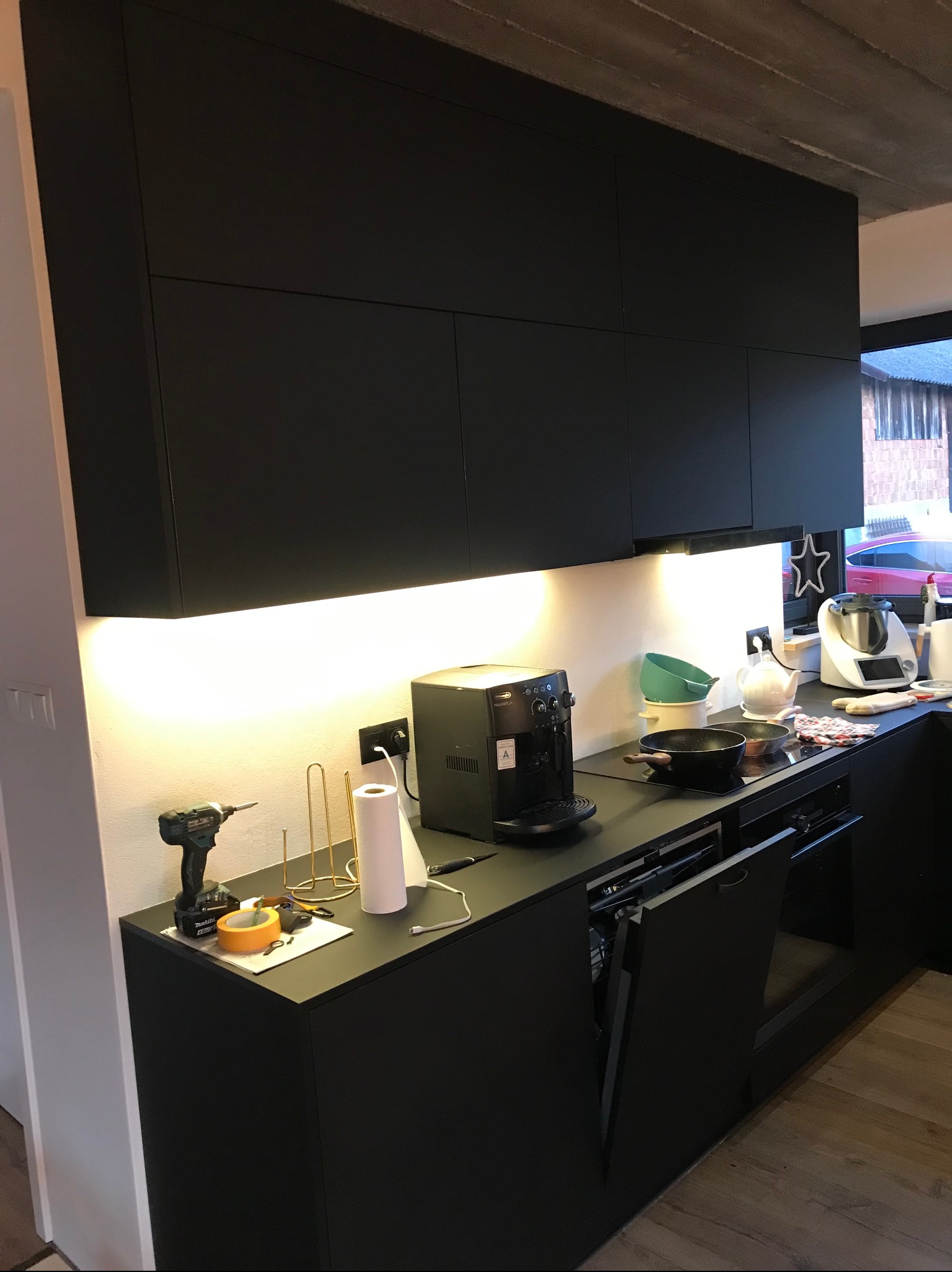 Nowoczesna, czarna zabudowa kuchenna z oświetleniem LED podszafkowym. Na blacie ekspres do kawy i akcesoria. Widoczna wkrętarka sugeruje etap montażu.