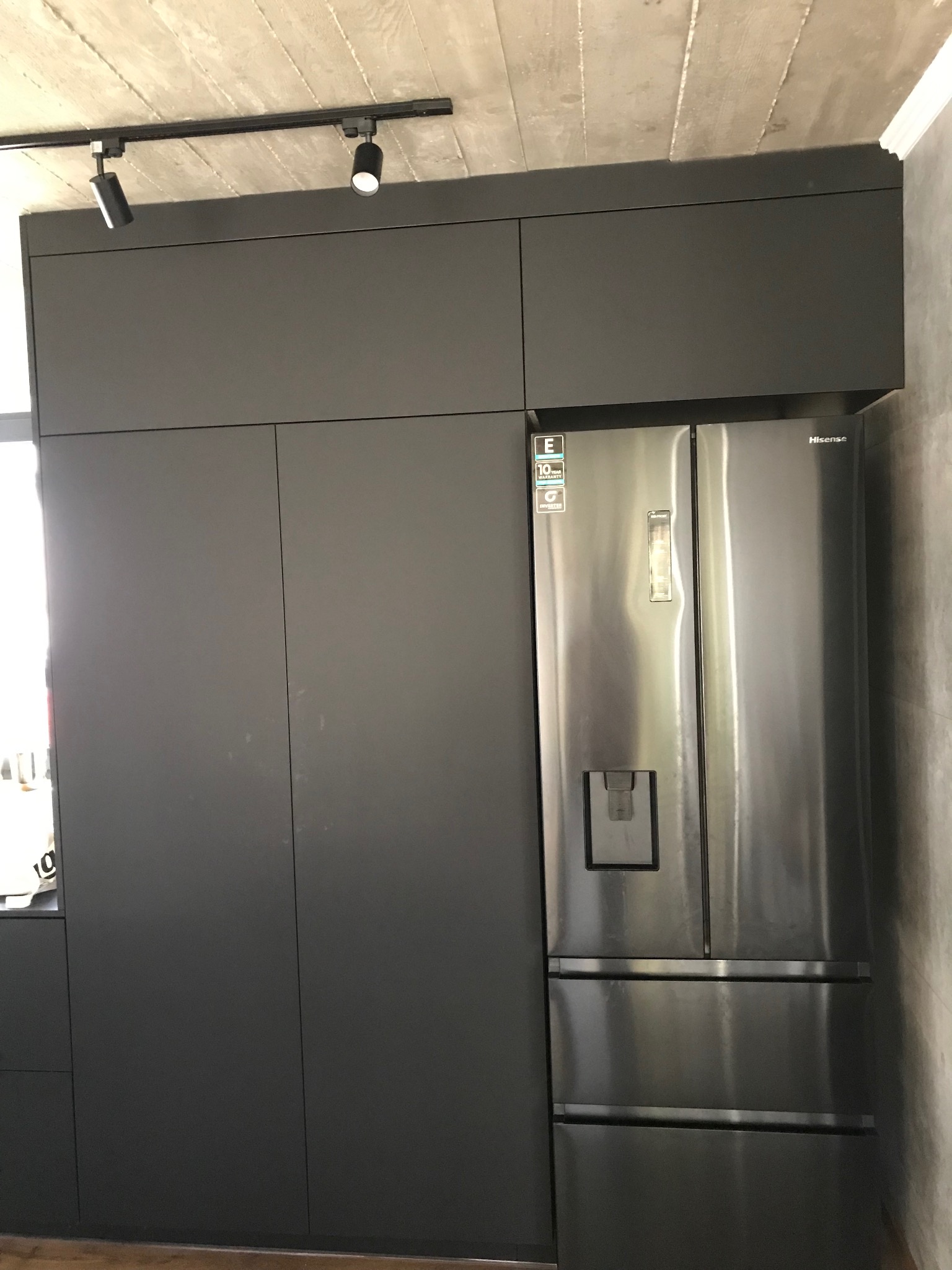 Nowoczesna kuchnia z zabudową w kolorze grafitu i dużą, dwudrzwiową lodówką Hisense. Minimalistyczny design, surowy beton na suficie i oświetlenie punktowe.
