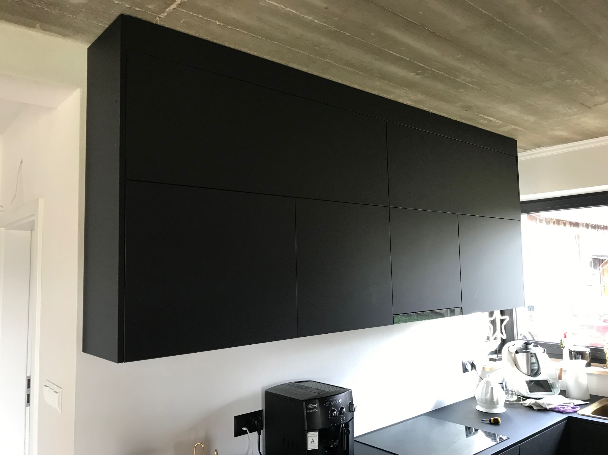 Czarne, minimalistyczne szafki kuchenne wiszące pod surowym betonowym sufitem. Na blacie ekspres do kawy. Nowoczesny design.