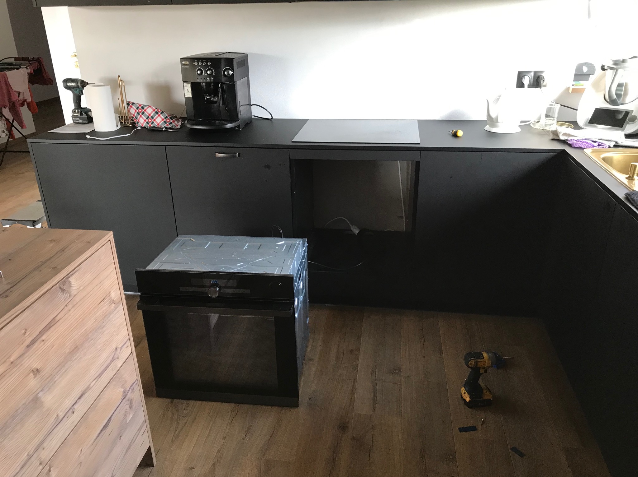Montaż piekarnika w czarnej zabudowie kuchennej. Na blacie ekspres do kawy, płyta indukcyjna i akcesoria. Na podłodze wkrętarka i elementy montażowe.