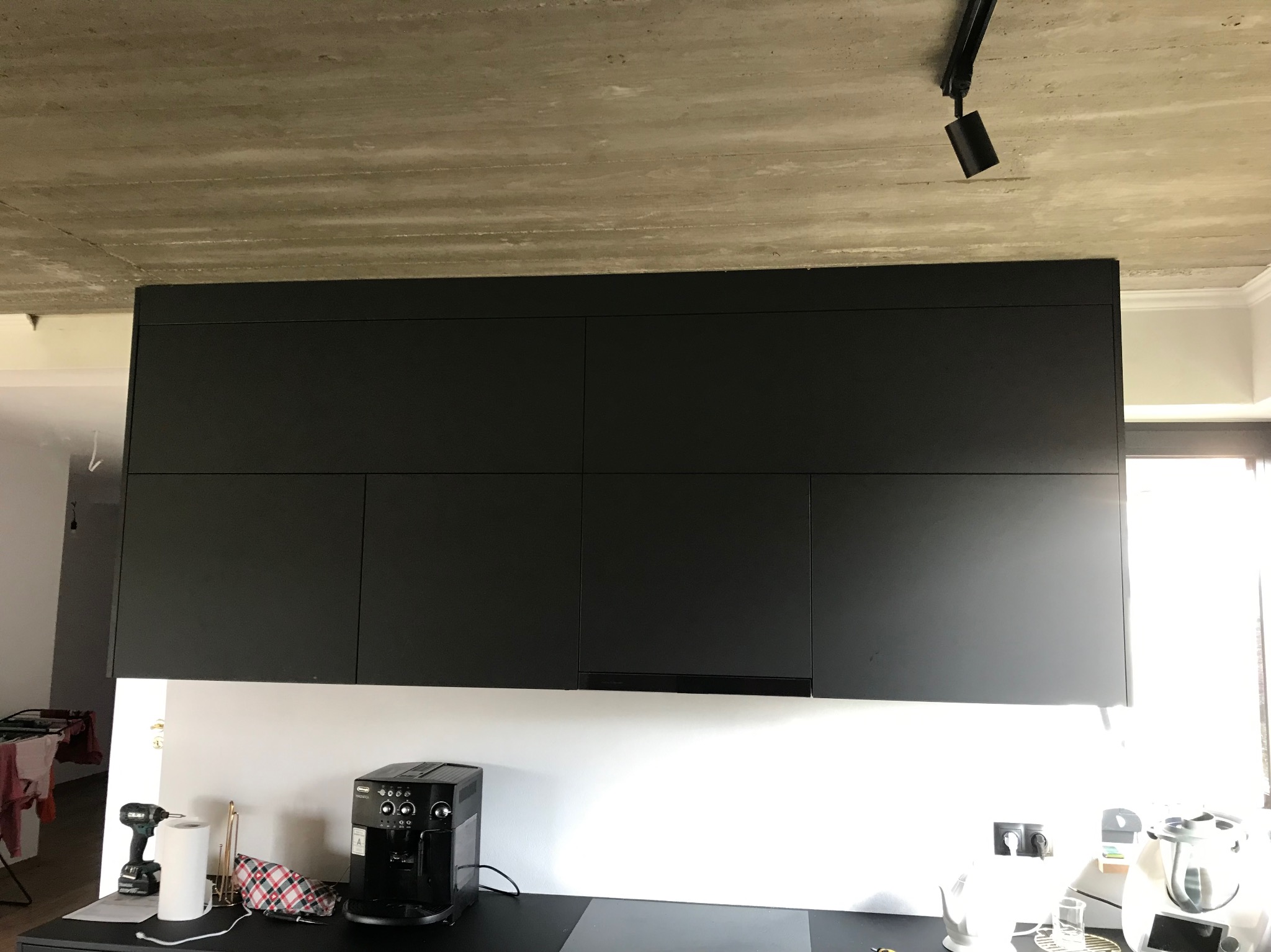 Czarne, matowe szafki kuchenne wiszące na białej ścianie, z widocznym betonowym sufitem i oświetleniem punktowym. Na blacie ekspres do kawy i narzędzia.