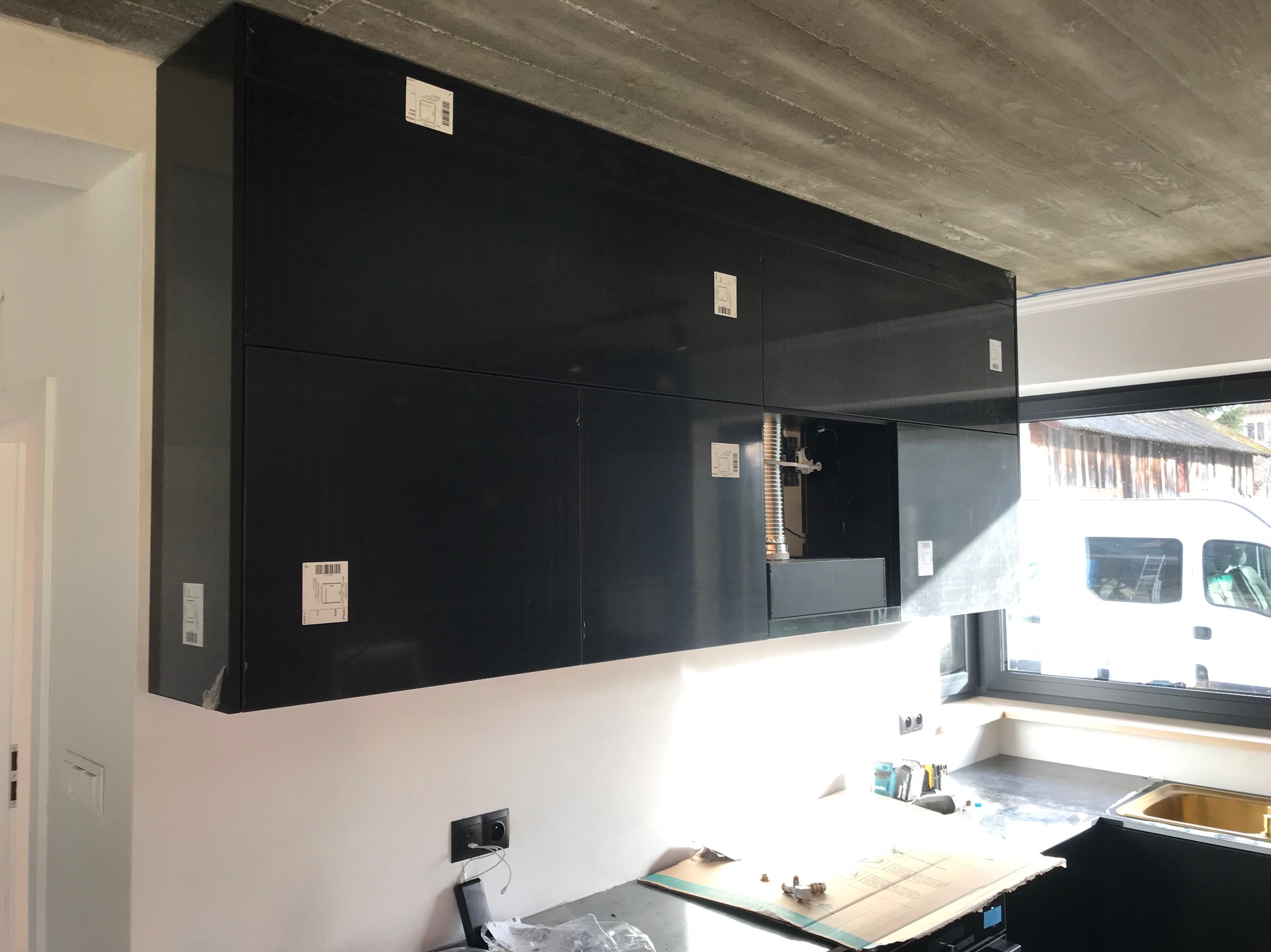 Częściowo zamontowane, czarne szafki kuchenne pod betonowym sufitem. Widoczne okno z białym busem na zewnątrz i elementy instalacji. Etap montażu kuchni na wymiar.
