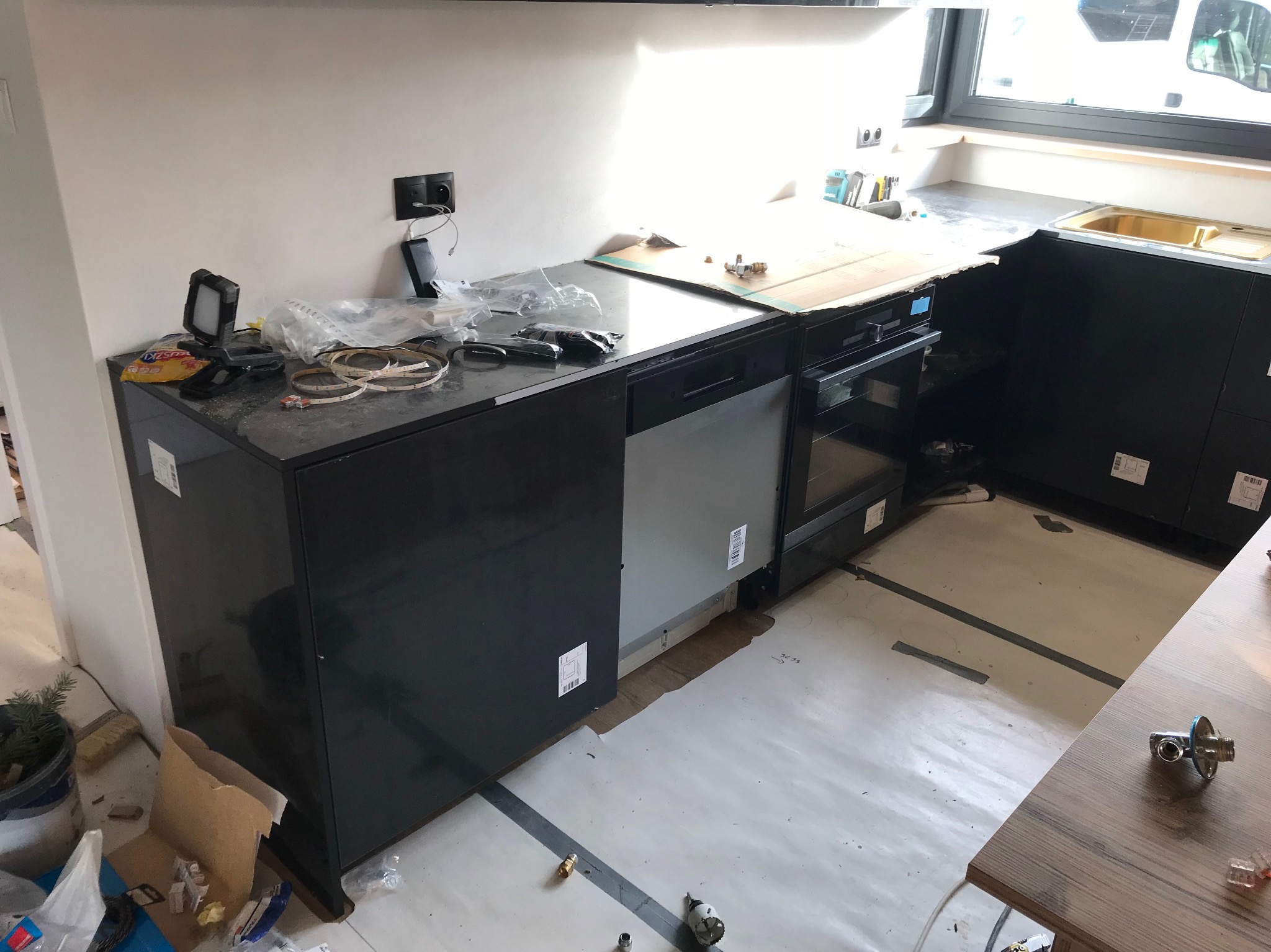 Częściowo zamontowana, nowoczesna, czarna zabudowa kuchenna z widocznymi elementami instalacji i narzędziami, sugerująca etap montażu. Widok na okno i fragment blatu.