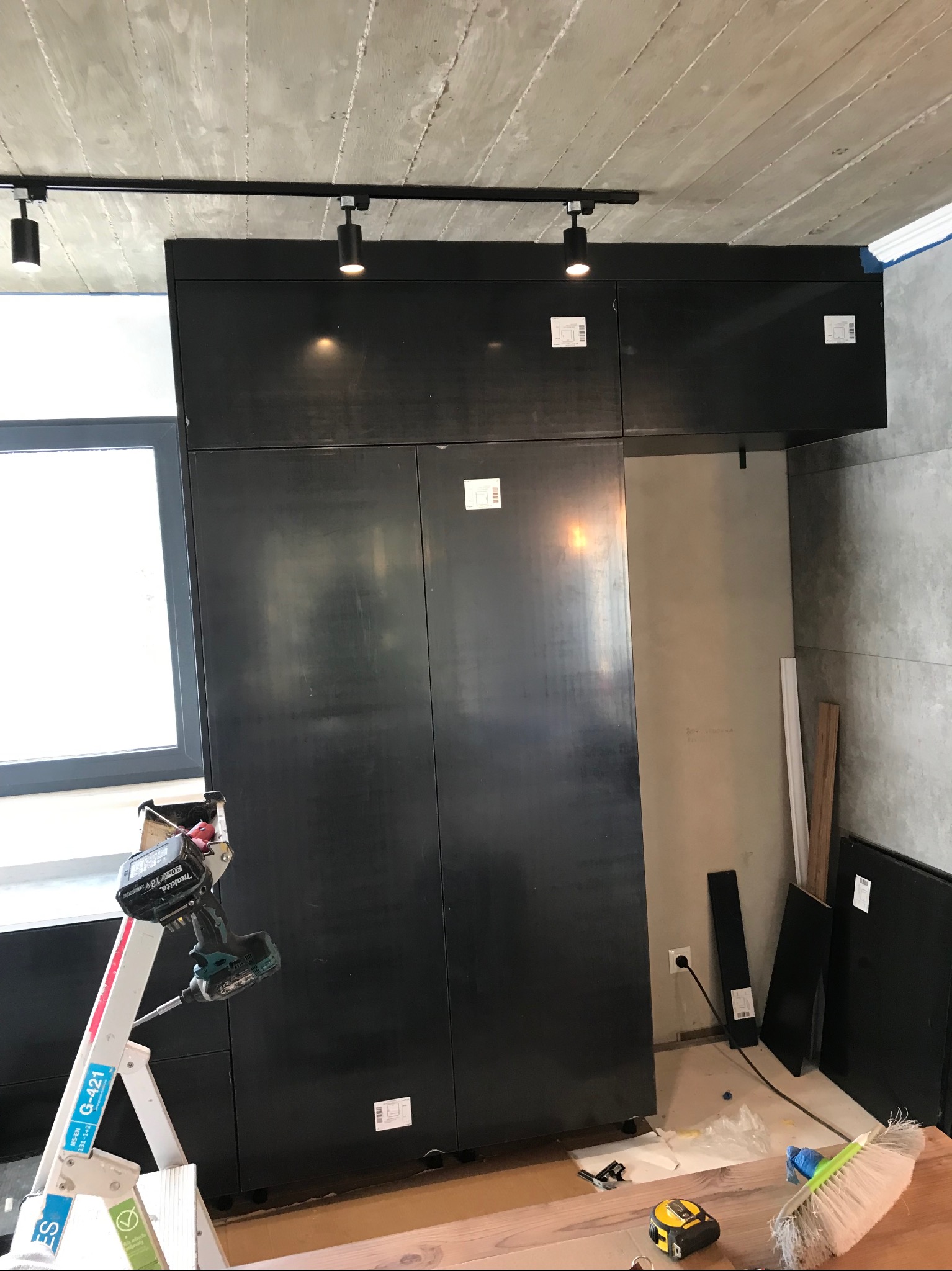 Montaż czarnej zabudowy kuchennej pod betonowym sufitem z oświetleniem punktowym. Widoczna drabina z wkrętarką i elementy wykończeniowe na podłodze.