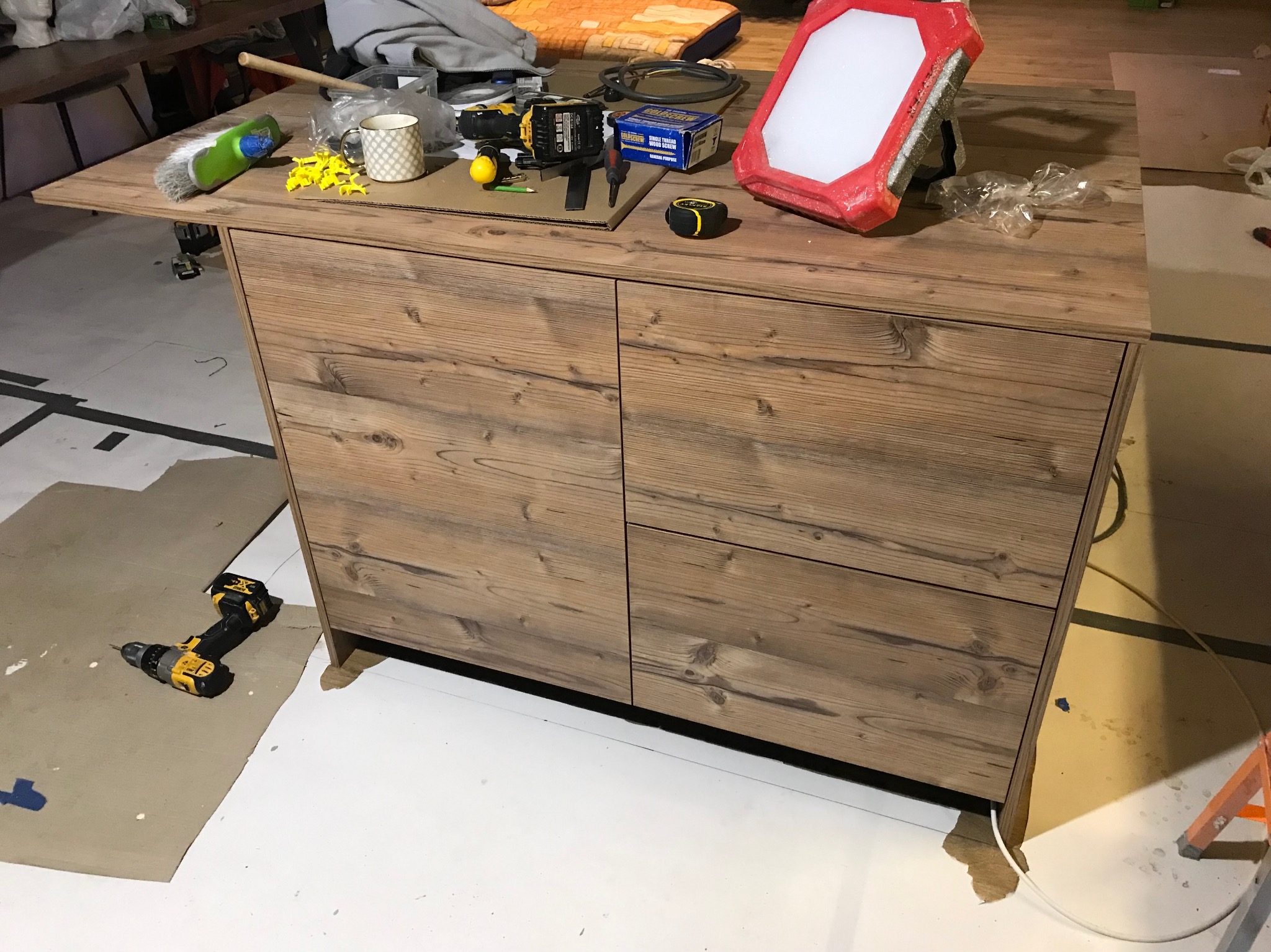 Montaż drewnianej szafki z szufladami, widoczne narzędzia: wiertarka, miarka, śrubokręt, lampa robocza. Na podłodze karton zabezpieczający. Ujęcie z bliska.