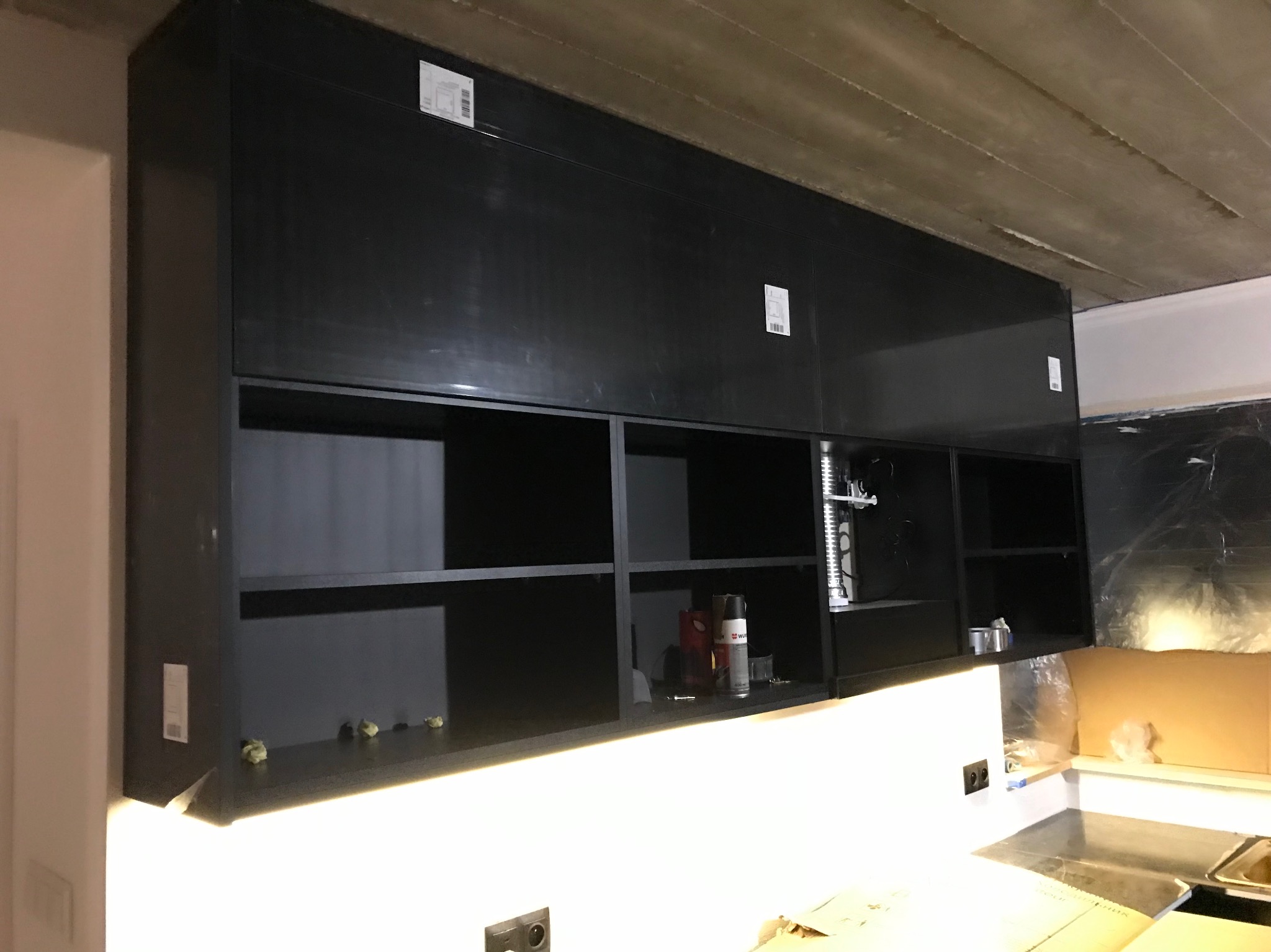 Czarne, wiszące szafki kuchenne bez frontów, z oświetleniem LED pod spodem, na tle surowego, betonowego sufitu. Widoczne akcesoria montażowe i zabezpieczenia.