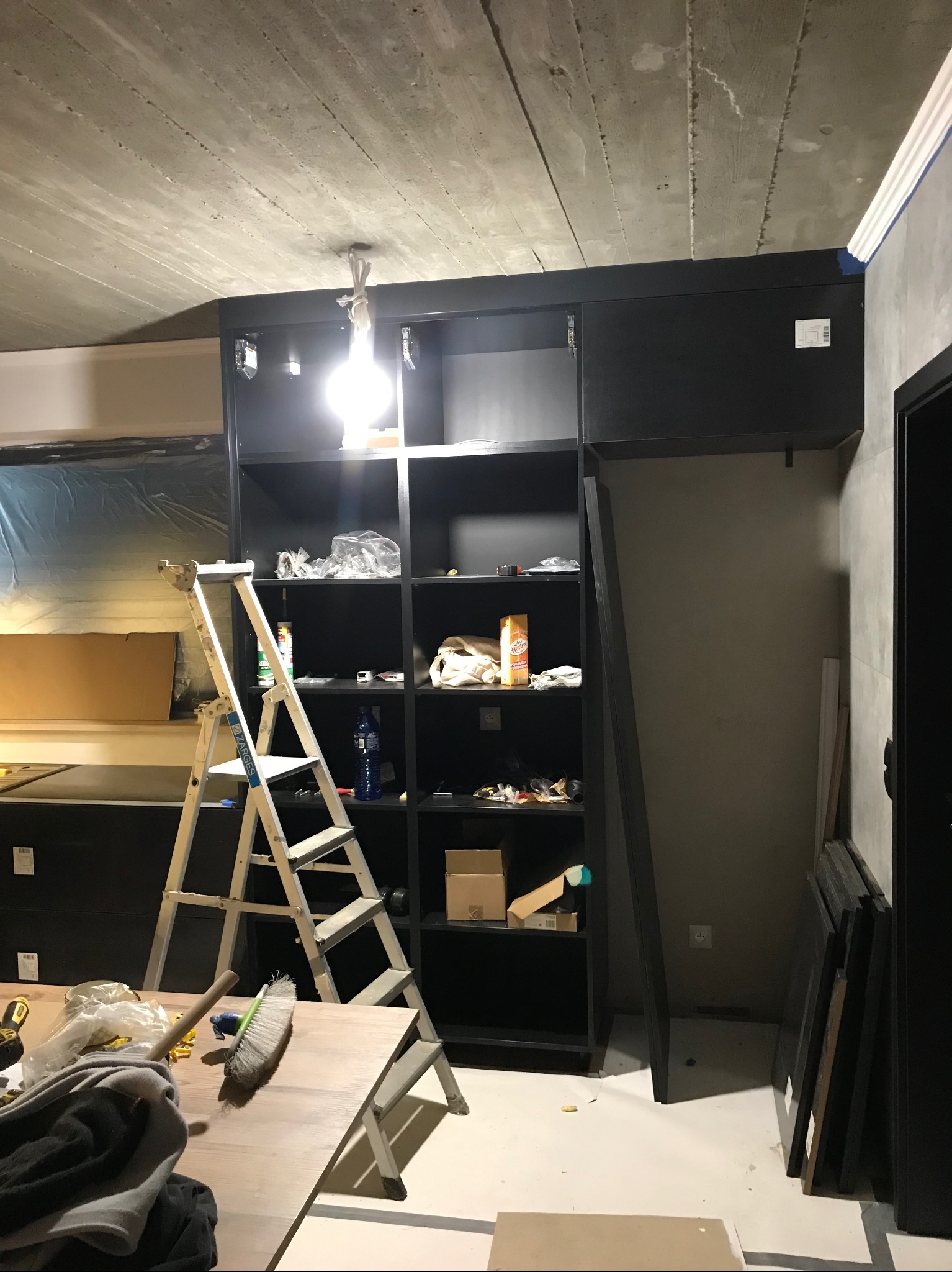 Ciemna zabudowa meblowa w trakcie montażu, widoczna drabina, narzędzia i materiały. Surowy betonowy sufit z widocznym oświetleniem. Etap prac wykończeniowych.