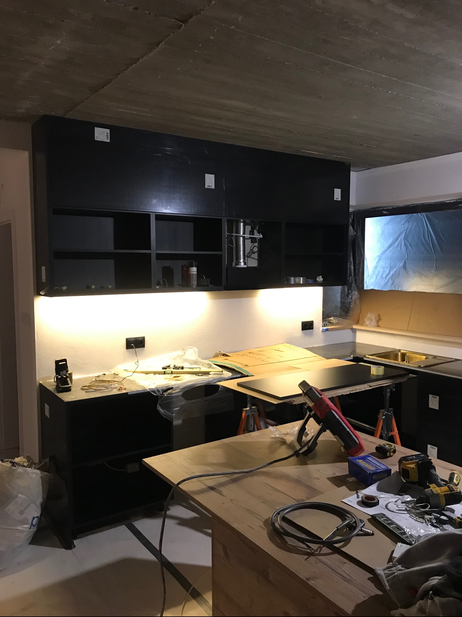 Częściowo zamontowana, czarna zabudowa kuchenna z oświetleniem LED, narzędzia na blacie, surowy betonowy sufit. Etap montażu kuchni na wymiar.