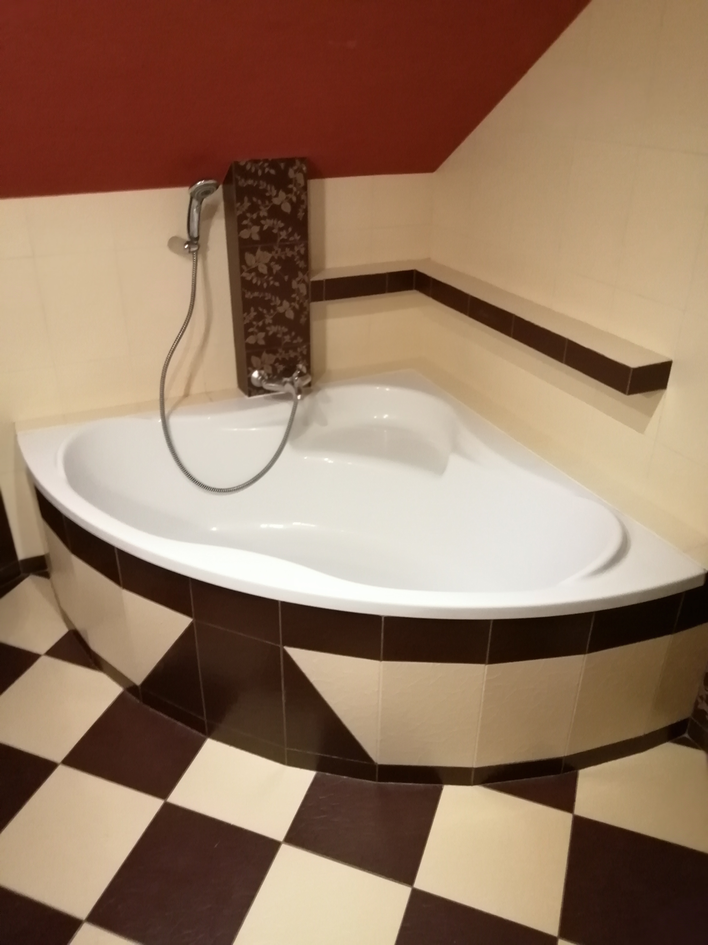 Narożna wanna z baterią i słuchawką prysznicową, obudowana płytkami w odcieniach brązu i beżu, z wzorem szachownicy na podłodze i półką wzdłuż ściany