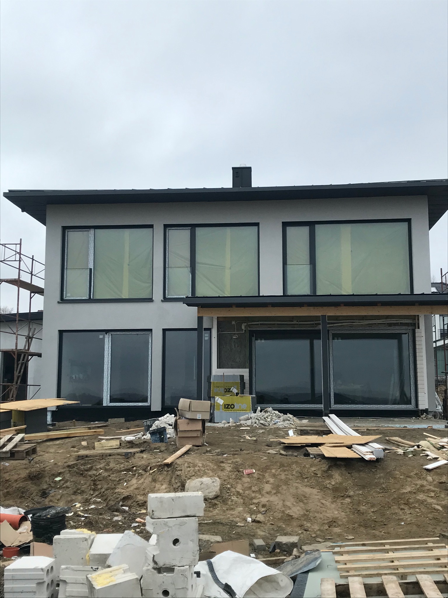 Surowy stan dwukondygnacyjnego domu z dużymi oknami, częściowo zabezpieczonymi folią, widoczne materiały budowlane i rusztowanie.