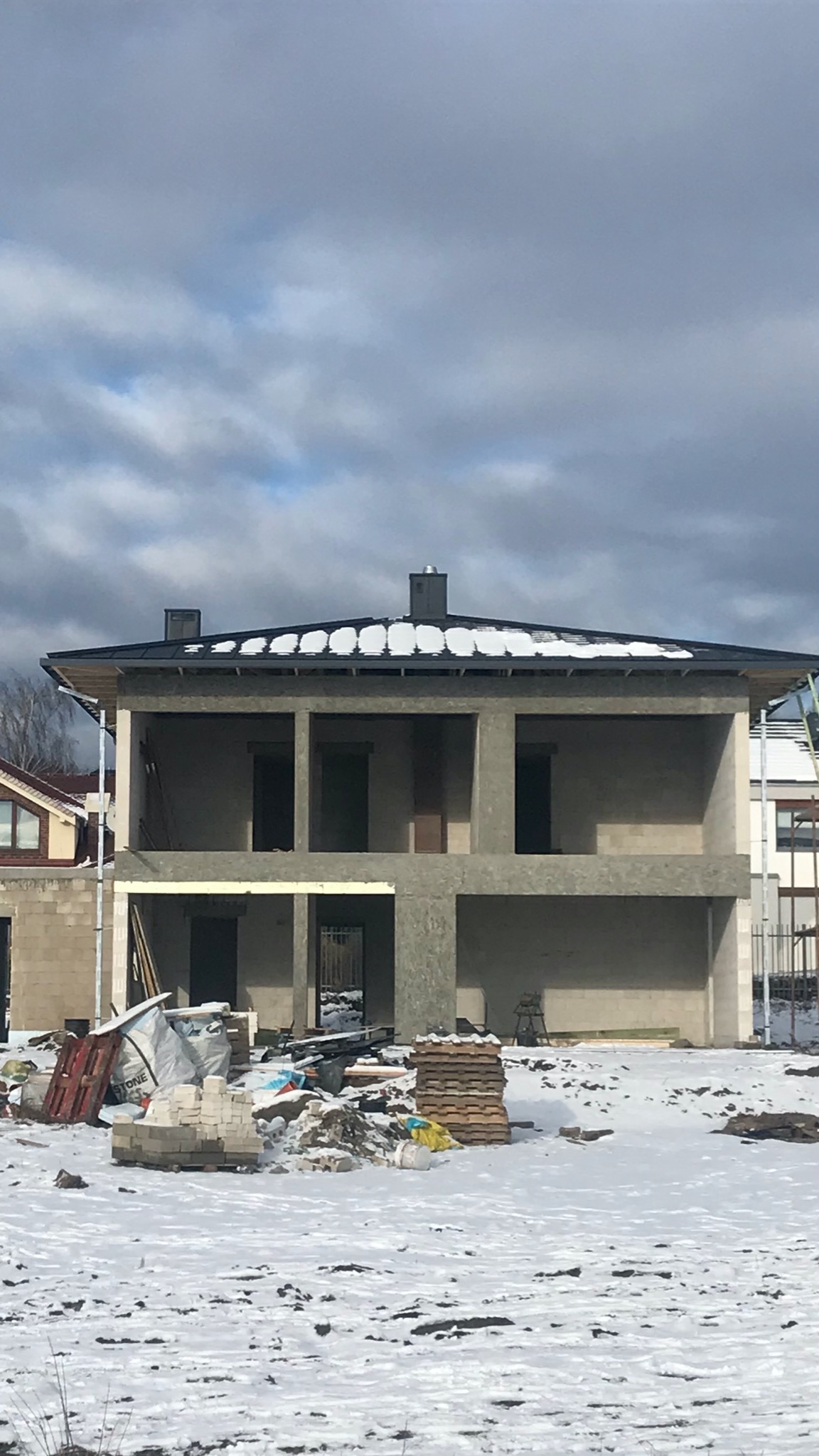Surowy stan dwukondygnacyjnego domu murowanego w trakcie budowy zimą, z widocznymi pustakami i śniegiem na dachu, z paletami i materiałami budowlanymi na ośnieżonym terenie budowy.