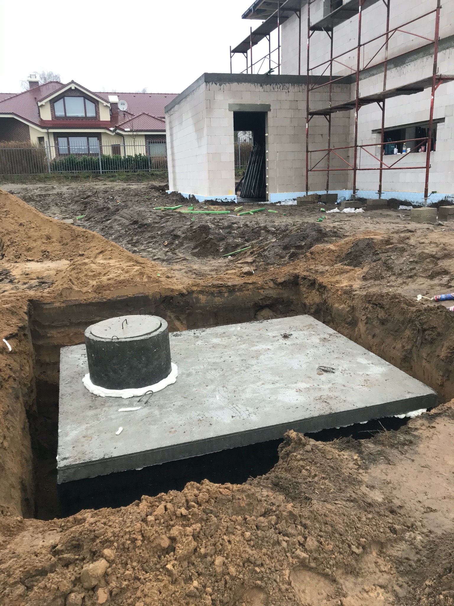 Betonowa płyta szamba umieszczona w wykopie, w tle budynek w stanie surowym otwartym z rusztowaniem i dom jednorodzinny.