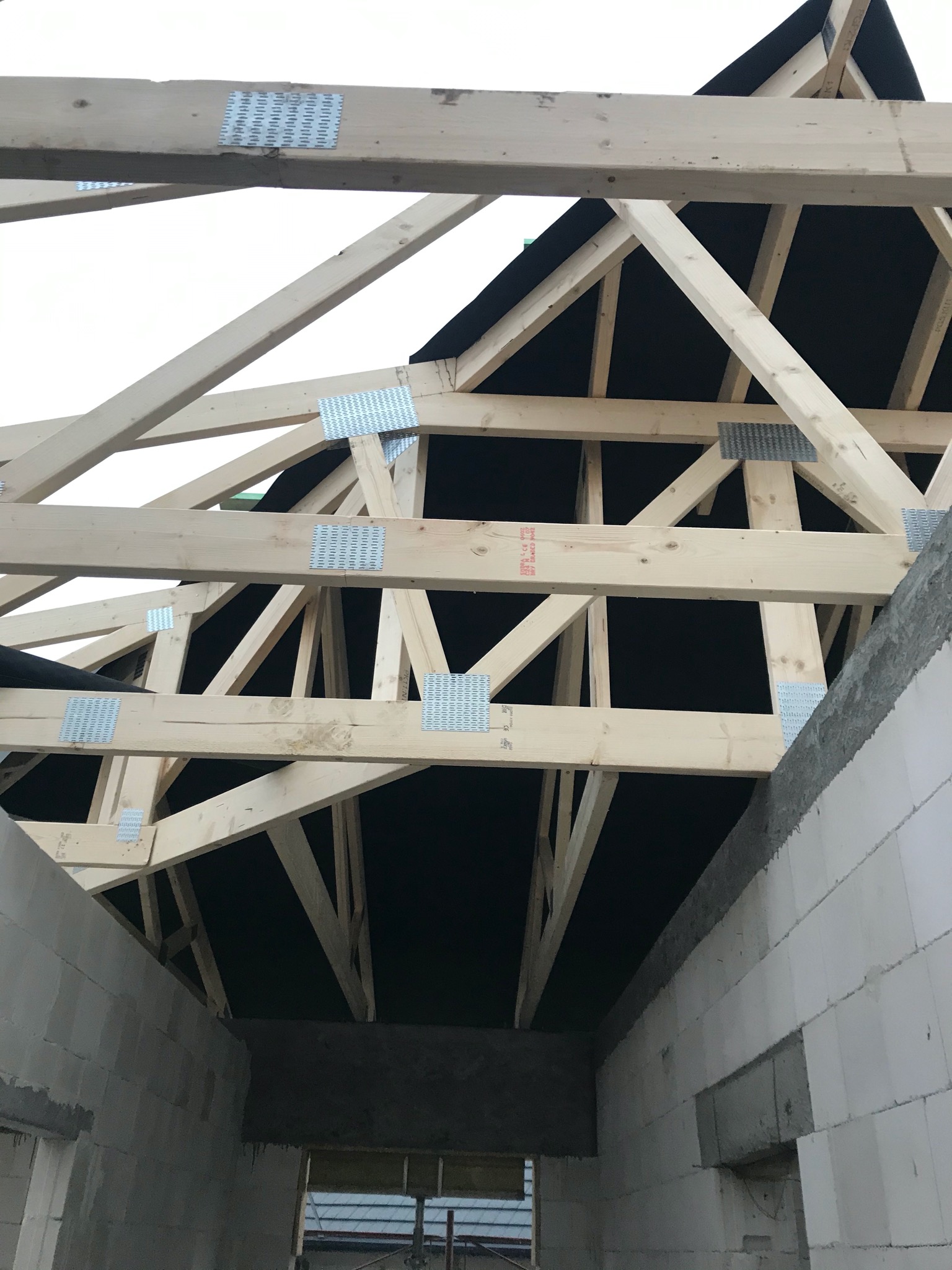 Konstrukcja drewniana dachu z widocznymi metalowymi łącznikami, czarna membrana dachowa i ściany z białych bloczków, budowa w trakcie realizacji.