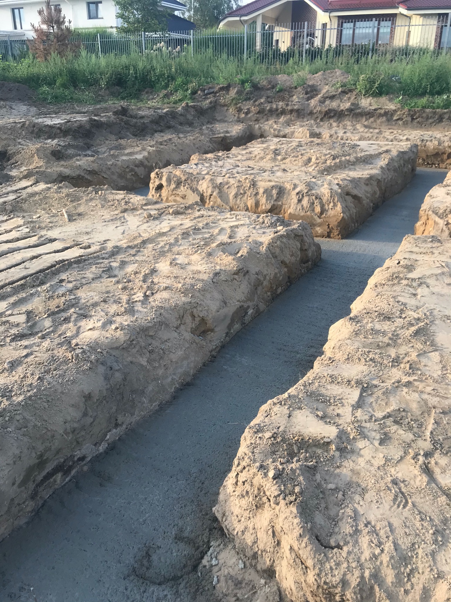 Wykopane i zalane betonem fundamenty pod budowę domu, widoczne ślady opon na ziemi i domy w tle.