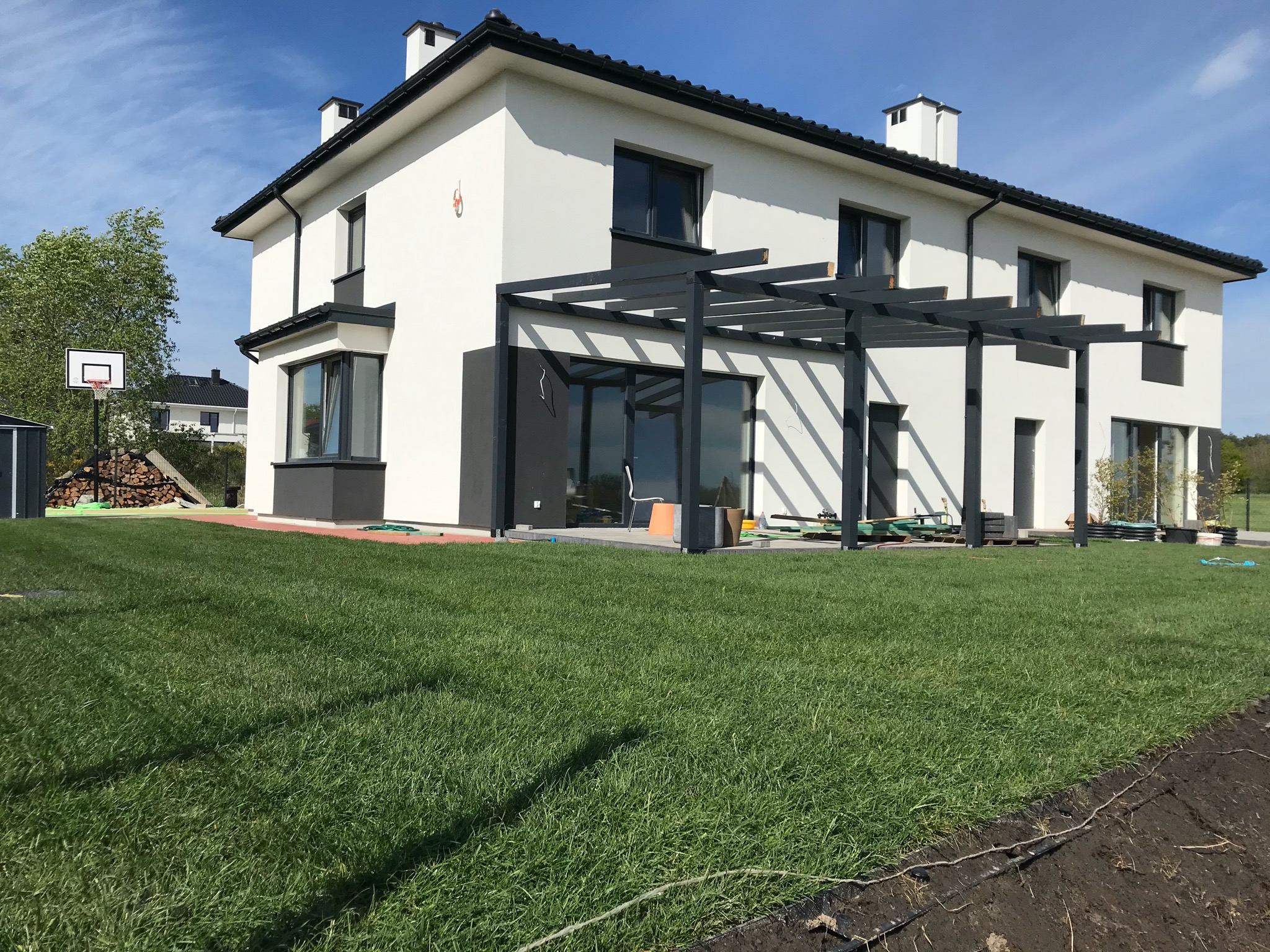 Nowoczesny dom z białymi ścianami i czarnym dachem, z pergolą nad tarasem, zadbanym trawnikiem i koszem do koszykówki w tle.