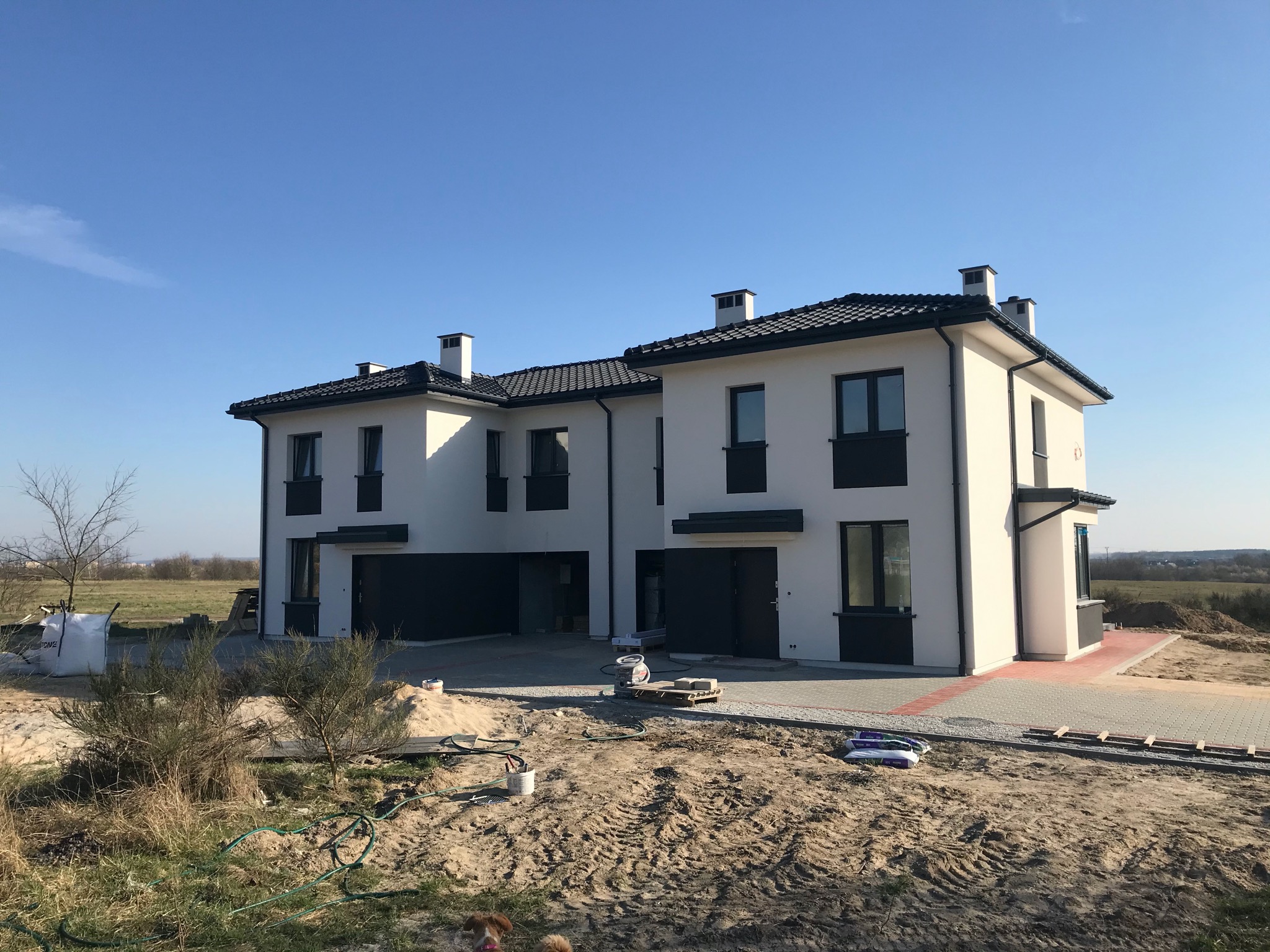 Dwa bliźniacze domy murowane w stanie surowym zamkniętym, z grafitową stolarką okienną i dachówką, na działce z widocznymi śladami budowy i niewielkim psem w rogu kadru.