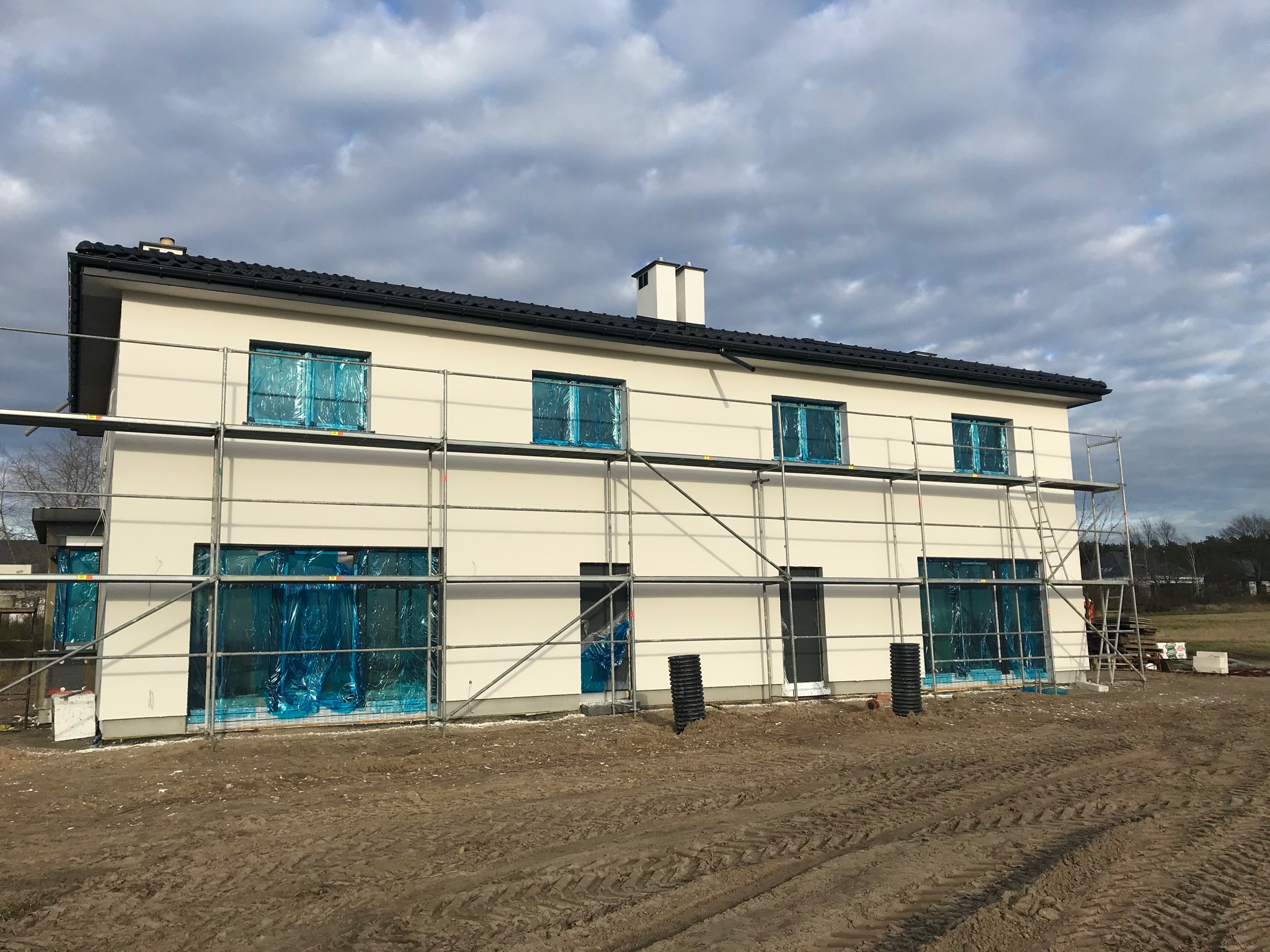Dwukondygnacyjny dom w trakcie budowy, z oknami zabezpieczonymi folią, otoczony rusztowaniami na tle pochmurnego nieba i przygotowanego terenu.