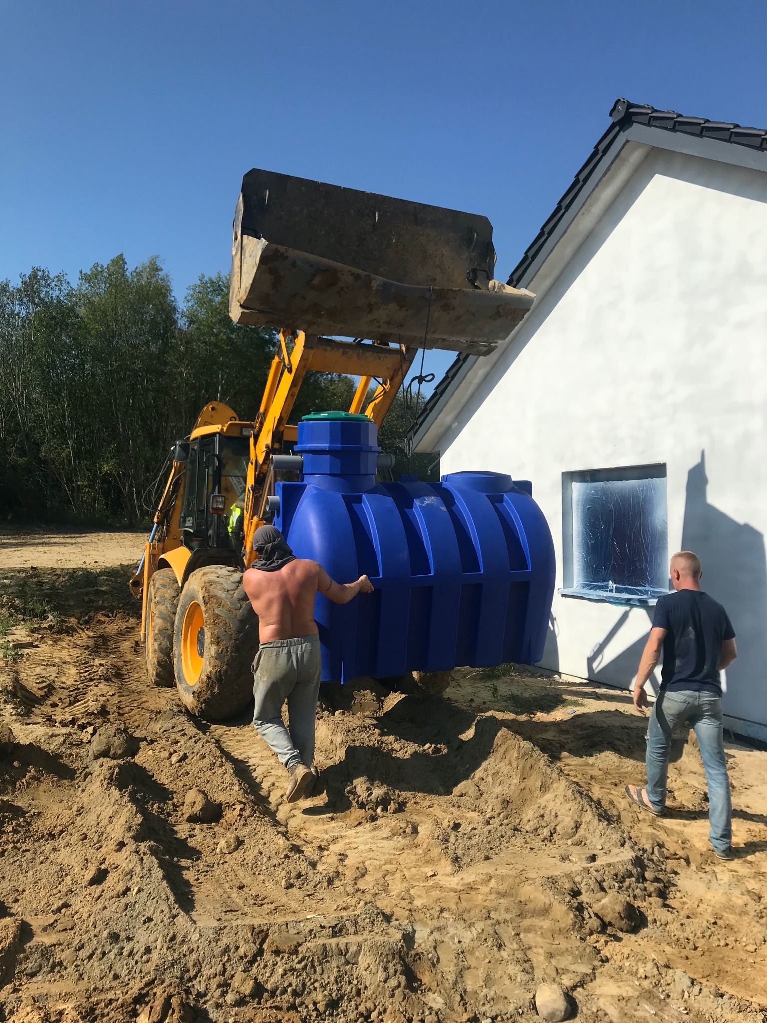 Żółta koparko-ładowarka transportuje niebieski zbiornik na wodę na placu budowy domu jednorodzinnego; pracownicy nadzorują proces, okno zabezpieczone folią.