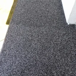 Szara, drobnoziarnista powierzchnia na balkonie, widoczna z góry, z fragmentem ściany i zieleni w tle.