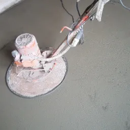 Szlifowanie świeżo wylanej posadzki za pomocą elektrycznej szlifierki tarczowej, widoczne ślady obróbki i niedoskonałości powierzchni w narożniku pomieszczenia.