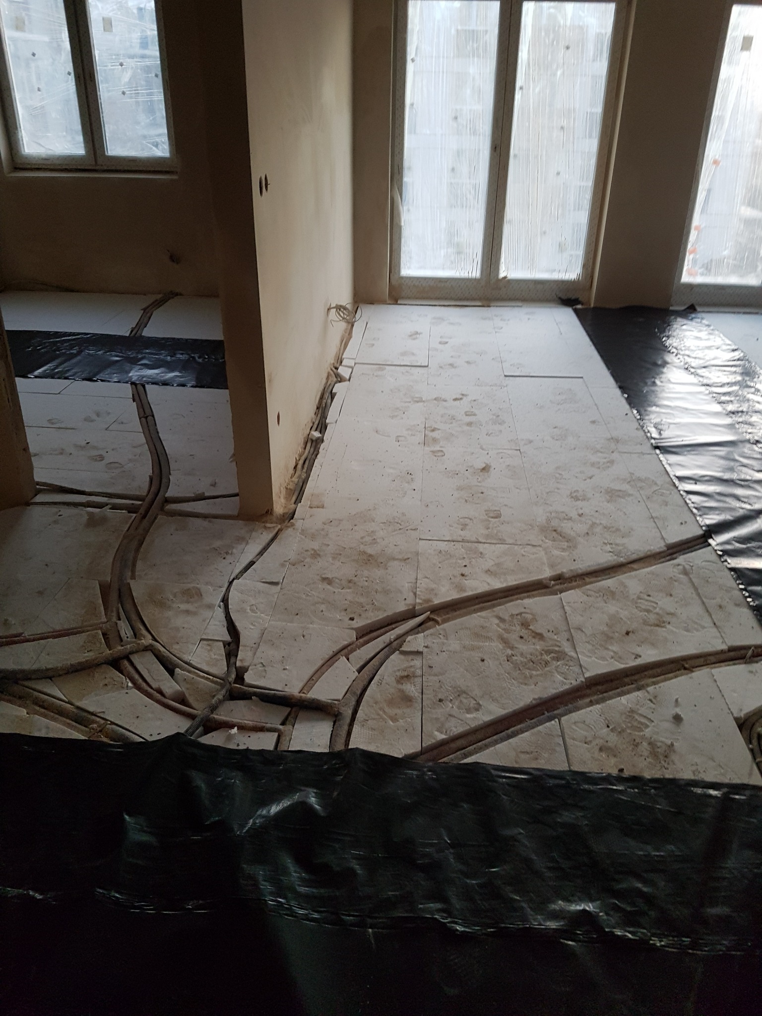 Przygotowanie podłoża pod posadzkę: widoczne rury ogrzewania podłogowego ułożone w wyżłobieniach w płytach izolacyjnych, zabezpieczone czarną folią, okna zakryte folią malarską.