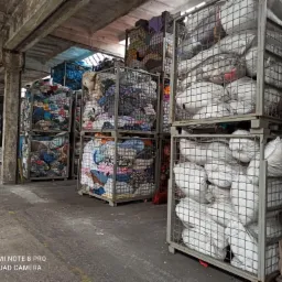 Magazyn z metalowymi koszami wypełnionymi workami i stertami różnorodnej odzieży, widoczne betonowe filary i posadzka.